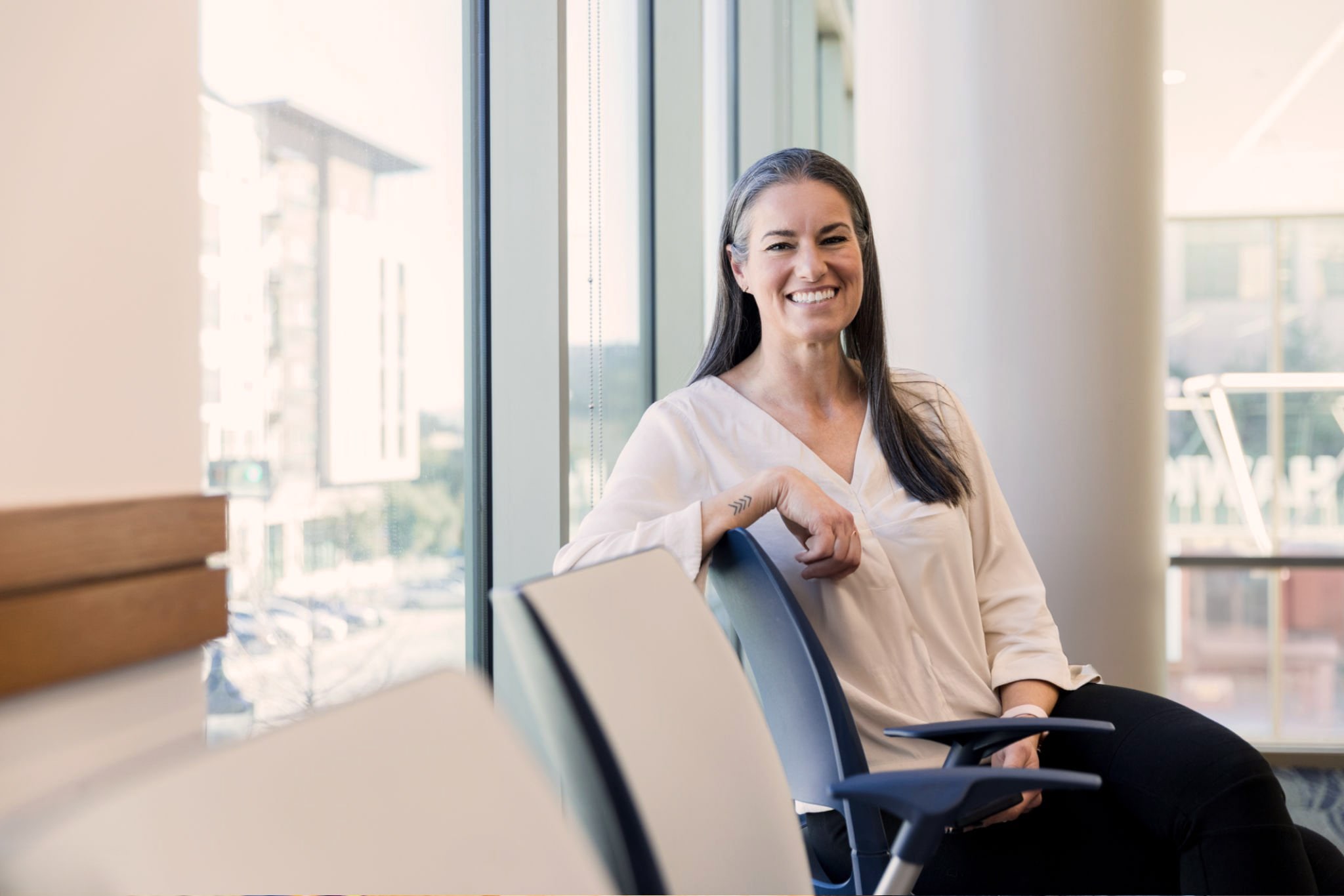 Smiling woman in a cream blouse seated on an office chair beside floor-to-ceiling windows