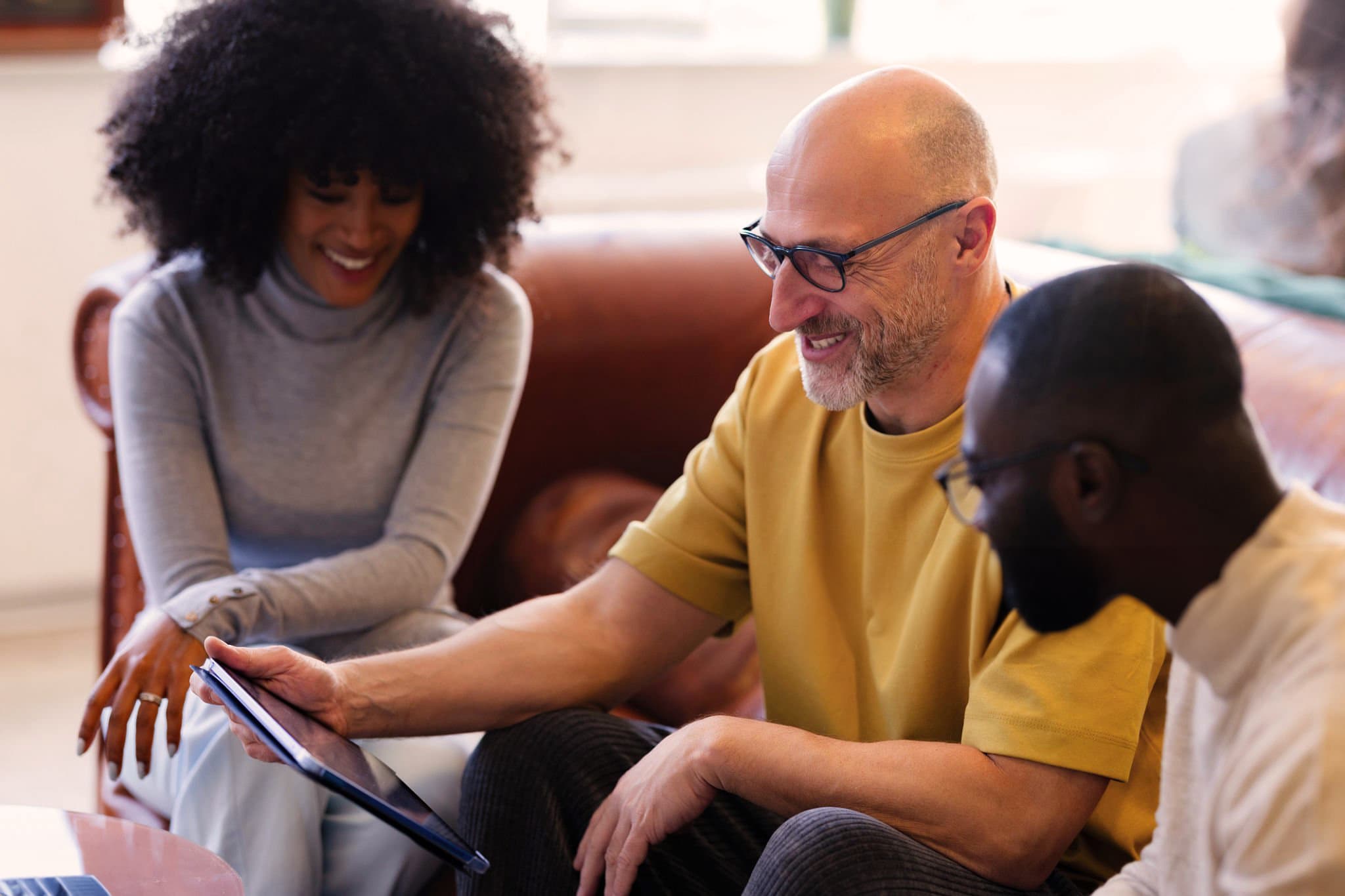 Three colleagues smiling while looking at a tablet together in a lounge area.