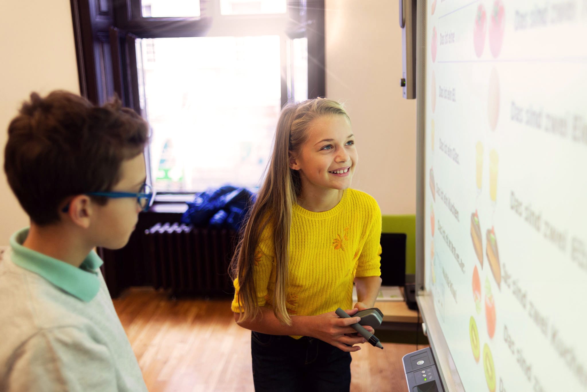 A pupil holding an interactive whiteboard pen smiles at the board while a classmate in glasses looks on