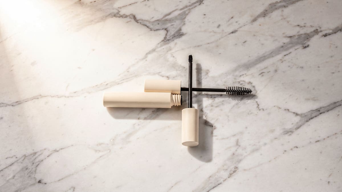 Light flat lay of a mascara tube and clean spoolie brush on white marble