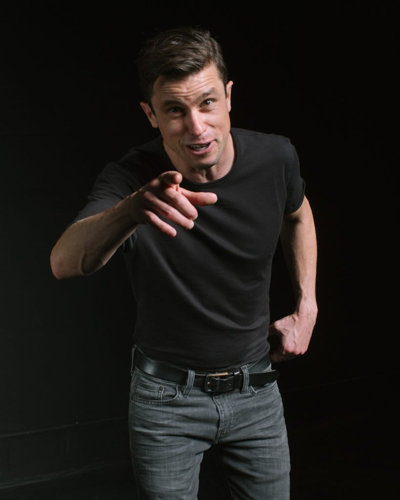 Dan Hoyle in a gray t-shirt pointing at the camera.