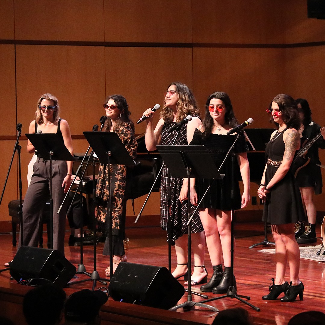 5 singers wearing sunglasses on stage with music stands and microphones