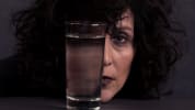 Actress with dark hair and dark smokey makeup peers from behind a water glass