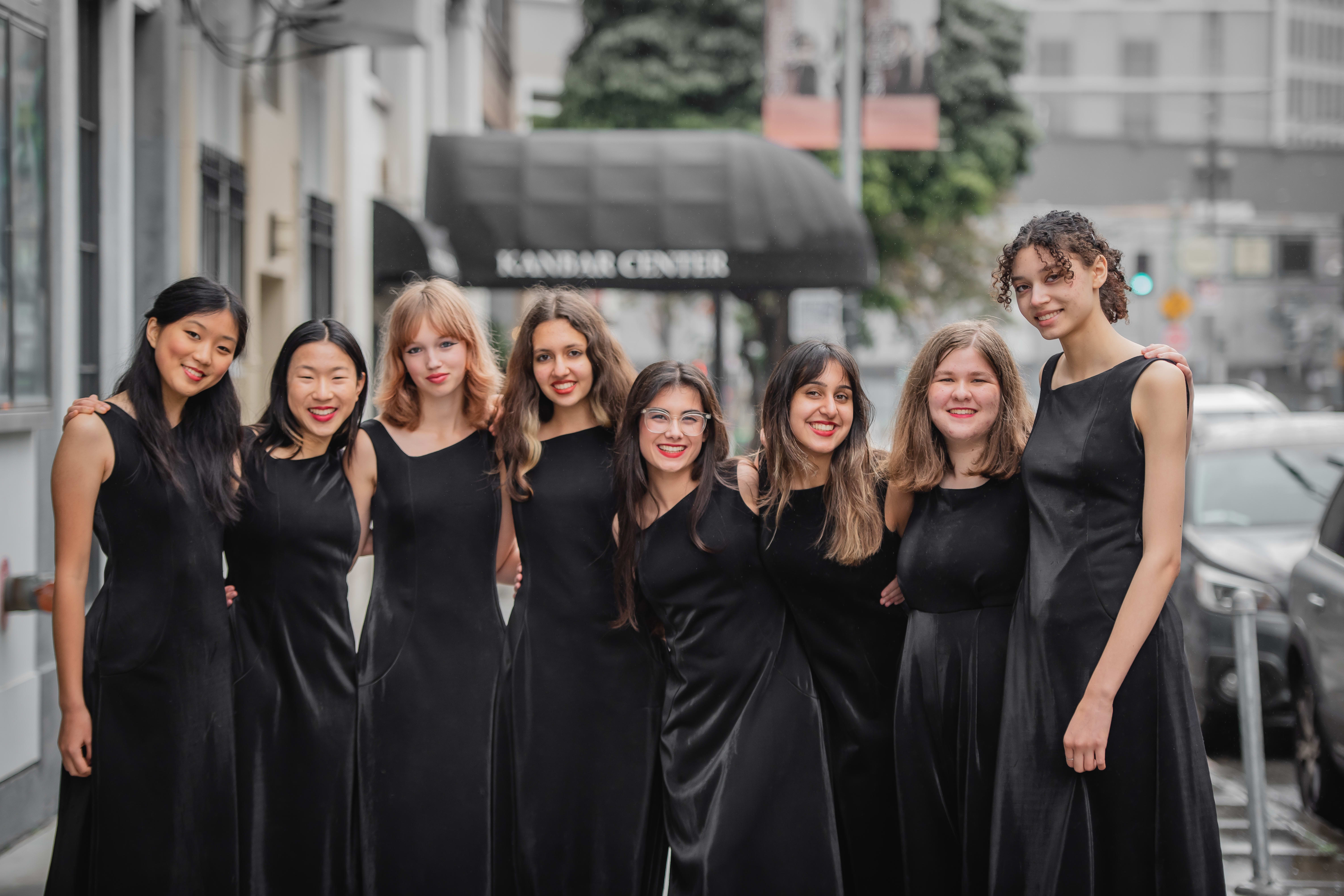 8 young singers in long black gowns smiling in a group