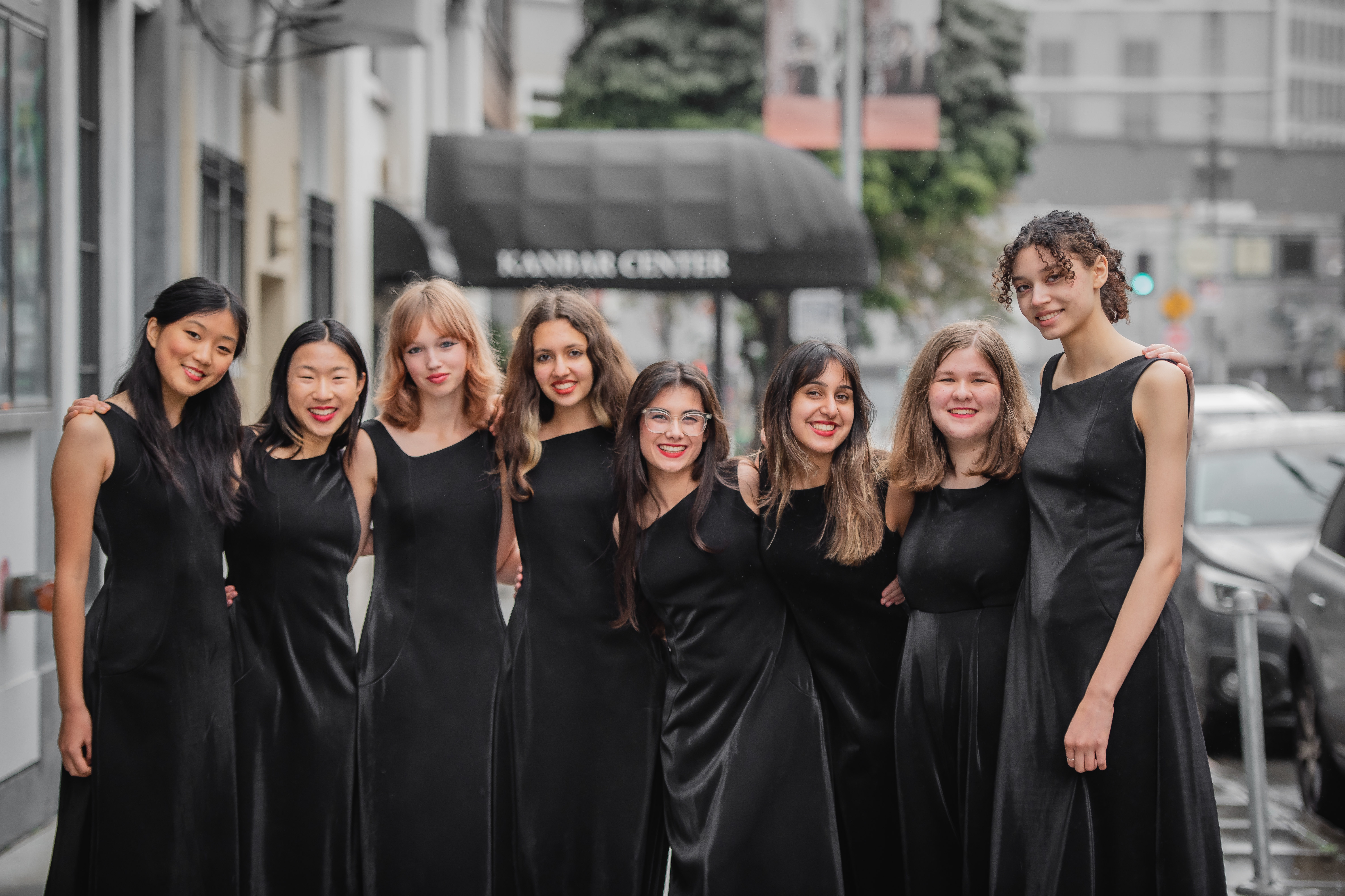 8 young singers in long black gowns smiling in a group