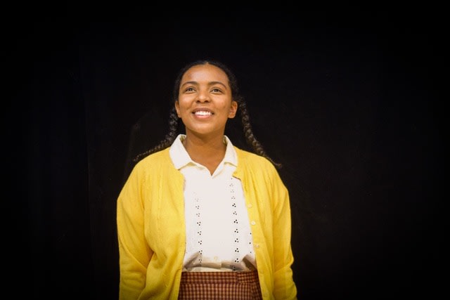 Black back drop, picture of Ernestine smiling hopefully in her yellow cardigan, white collared shirt, and mustard and maroon