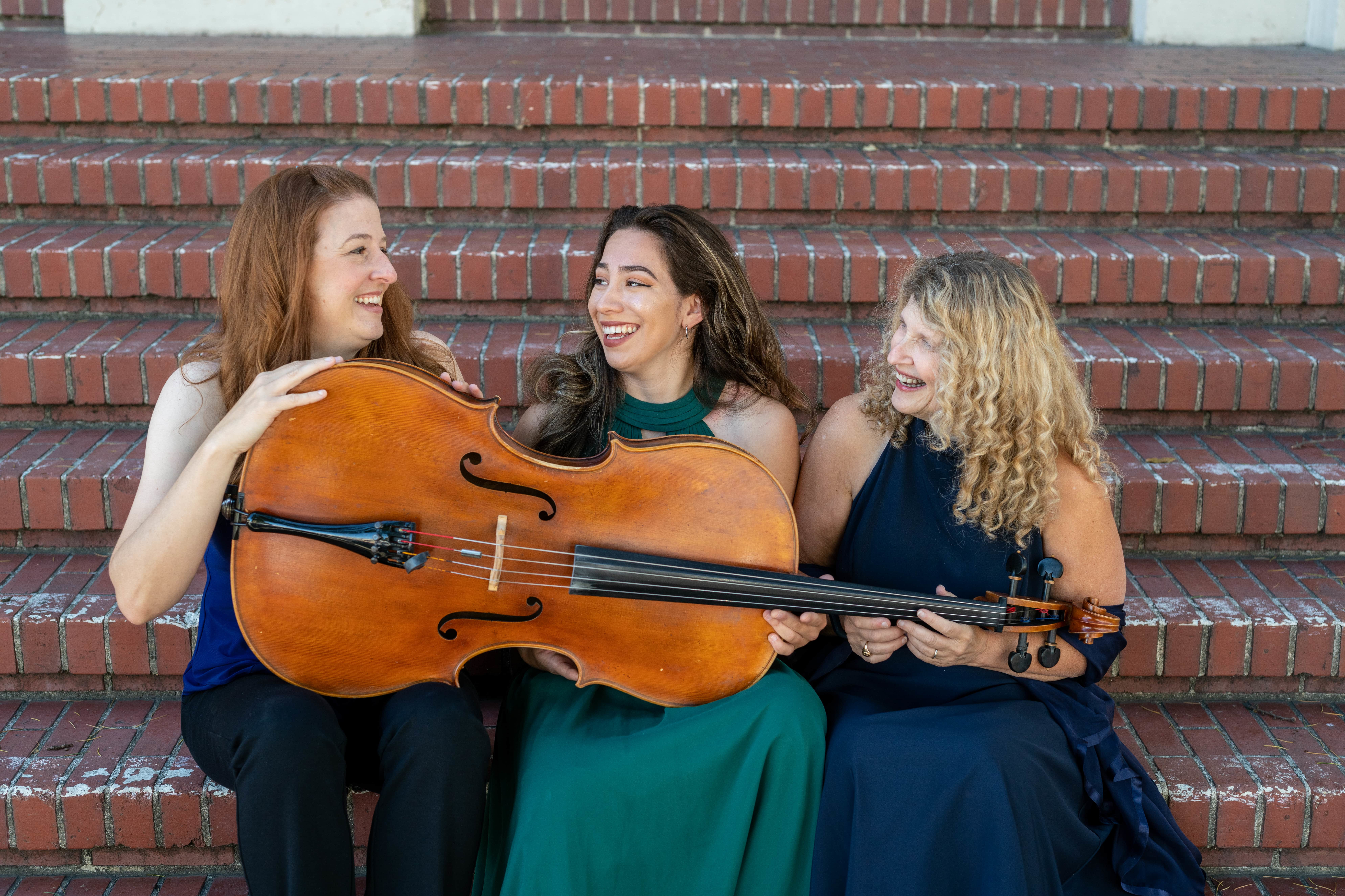 E4TT's members, Margaret Halbig, Abigail Monroe, Nanette McGuinness (l to r) setting on brick steps behind a cello.