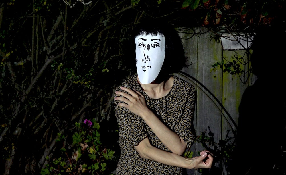 One artist standing in dark is wearing a oval-shaped paper mask on which a face is drawn with black ink.