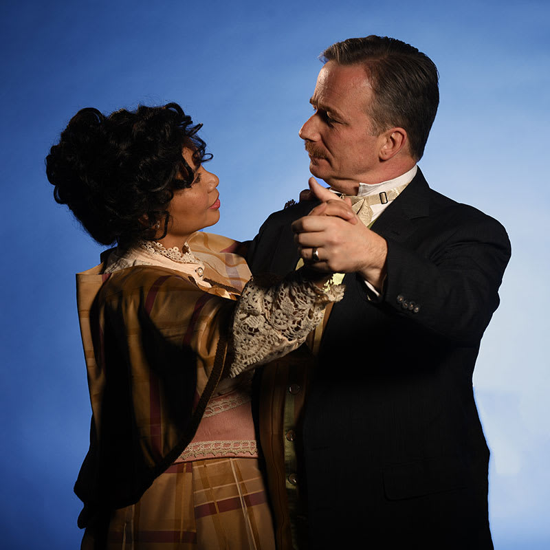 Michal Rhone and Jennifer Cuevas waltz together against a blue backdrop, dressed as characters from the Stephen Sondheim musi
