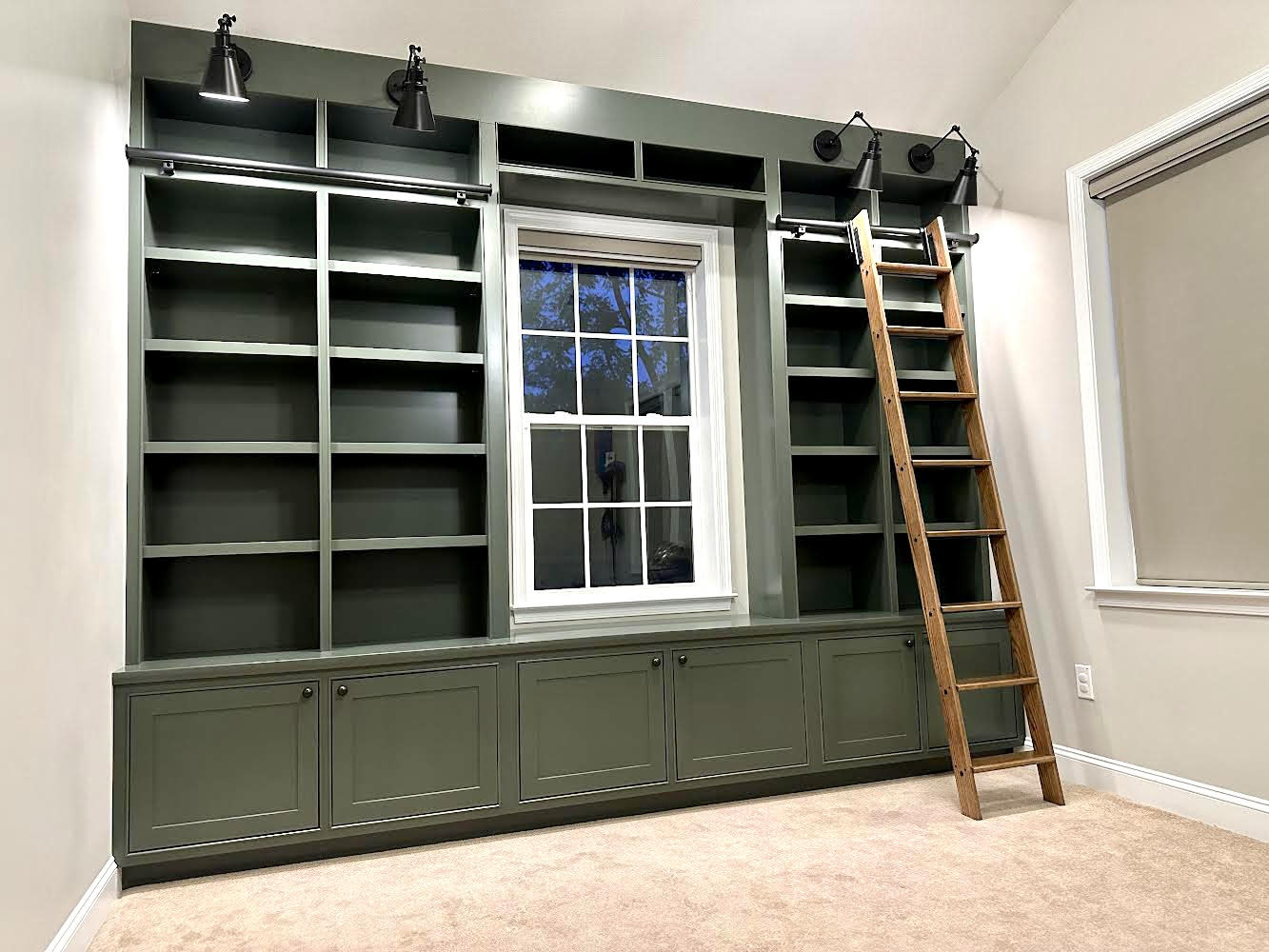 A large, custom-built library wall unit painted in a deep sage green, featuring numerous open bookshelves, cabinets