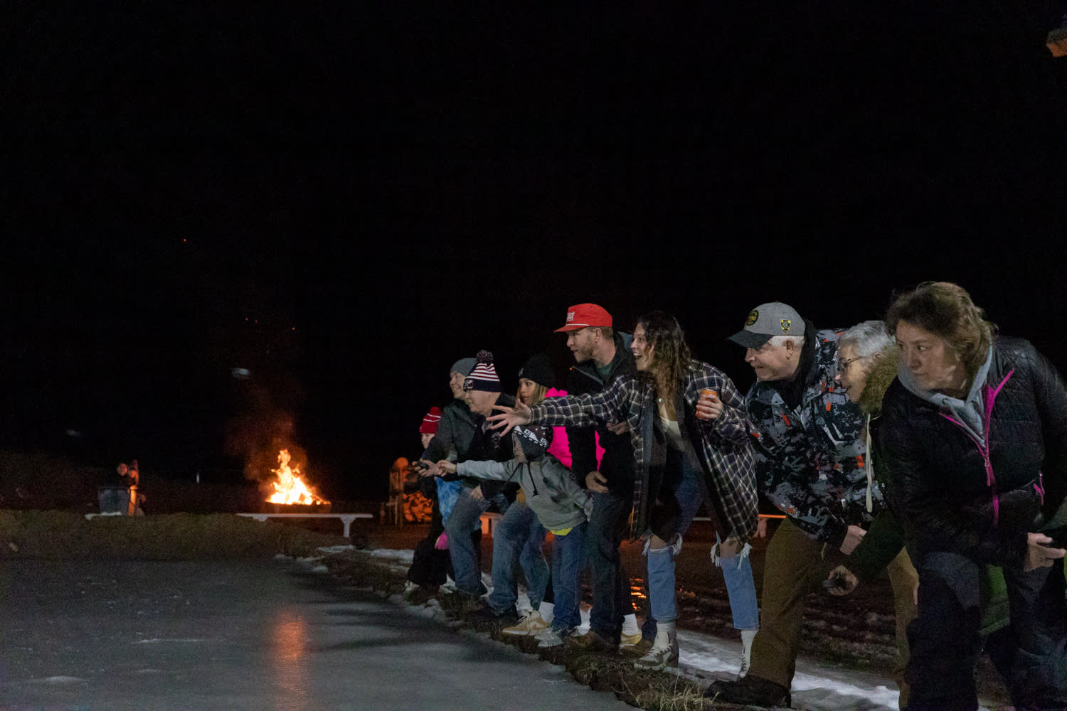 People lined up next to each other throwing pucks onto ice with a bonfire in the background at night time