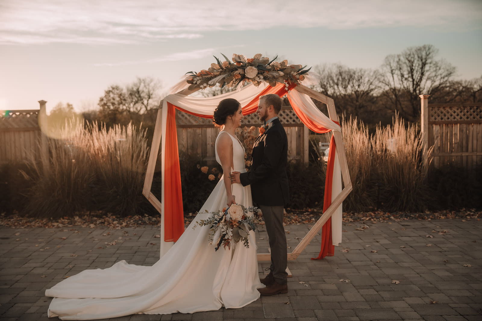 Bride and groom standing under a geometric arch with Sola Wood florals and draped fabric at sunset.