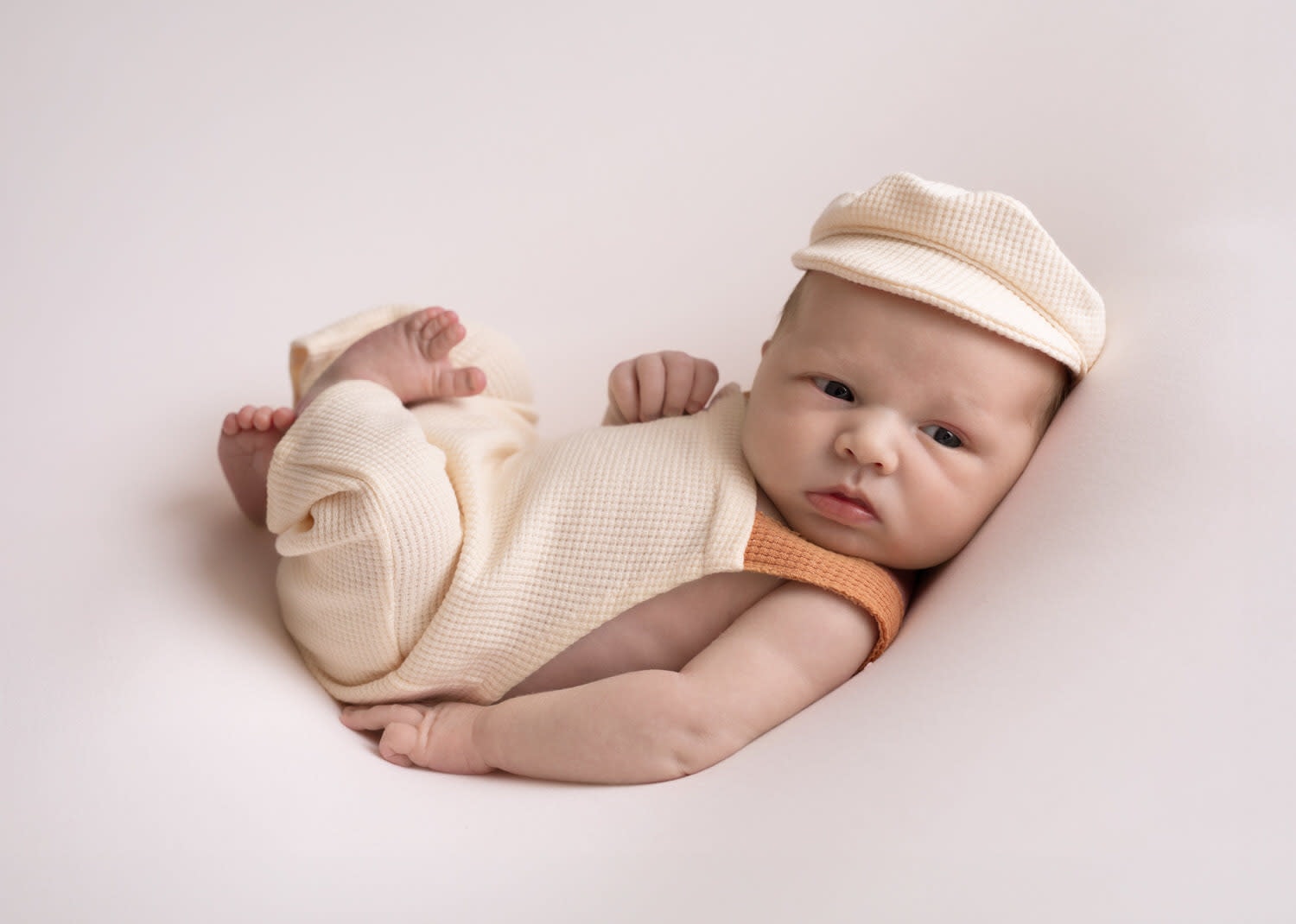 Newborn Boy in cream overalls, matching newsboy hat. On his back, feet up. Huck Finn Pose