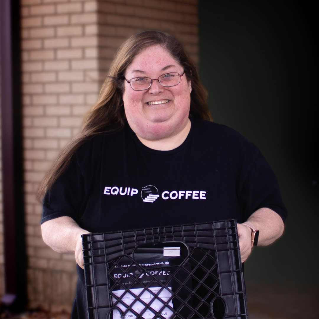 Picture of woman smiling, holding crate full of coffee. Dressed in a black tee shirt that says Equip Coffee