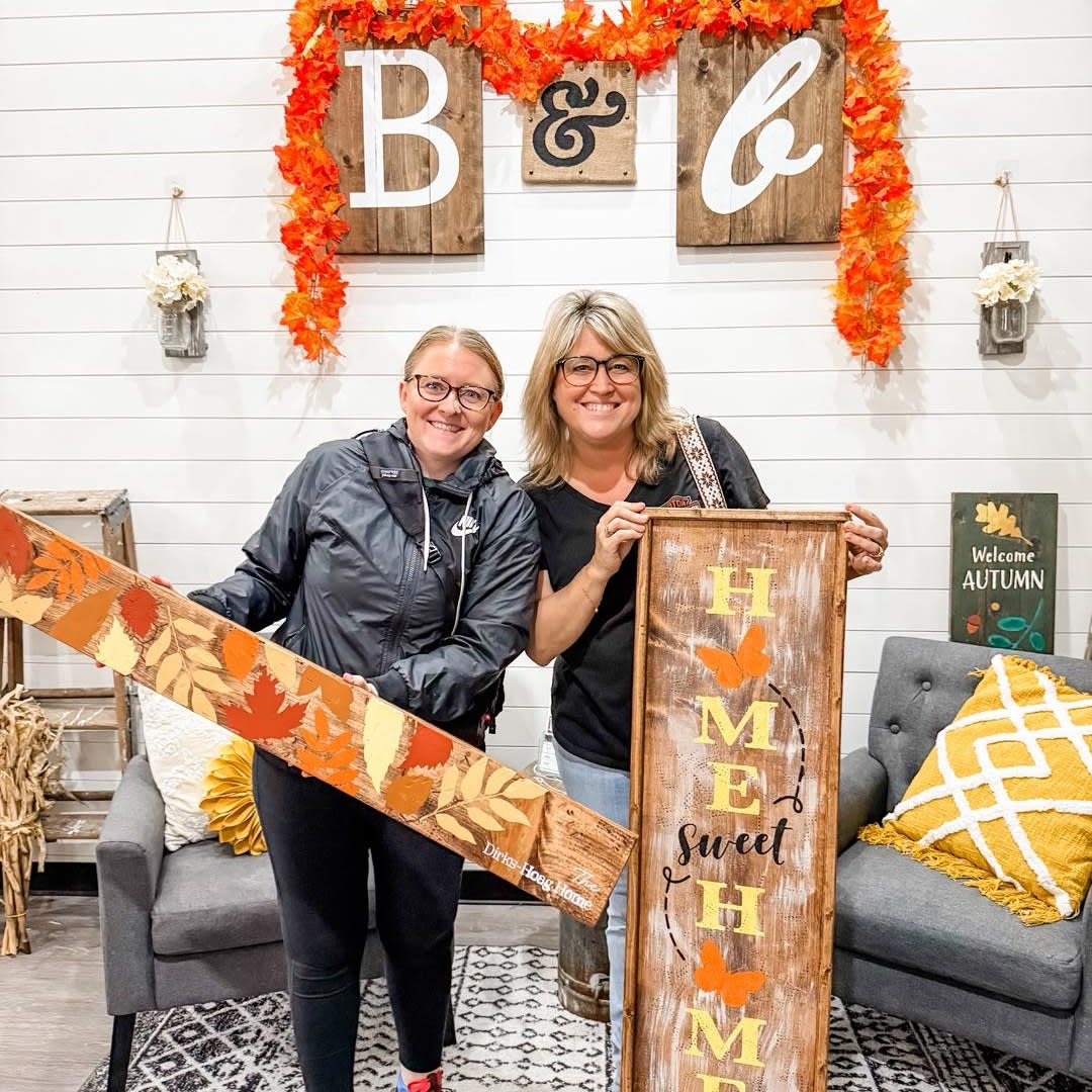 Two blonde women stand side by side displaying their painted wood signs made at a Board & Brush workshop.