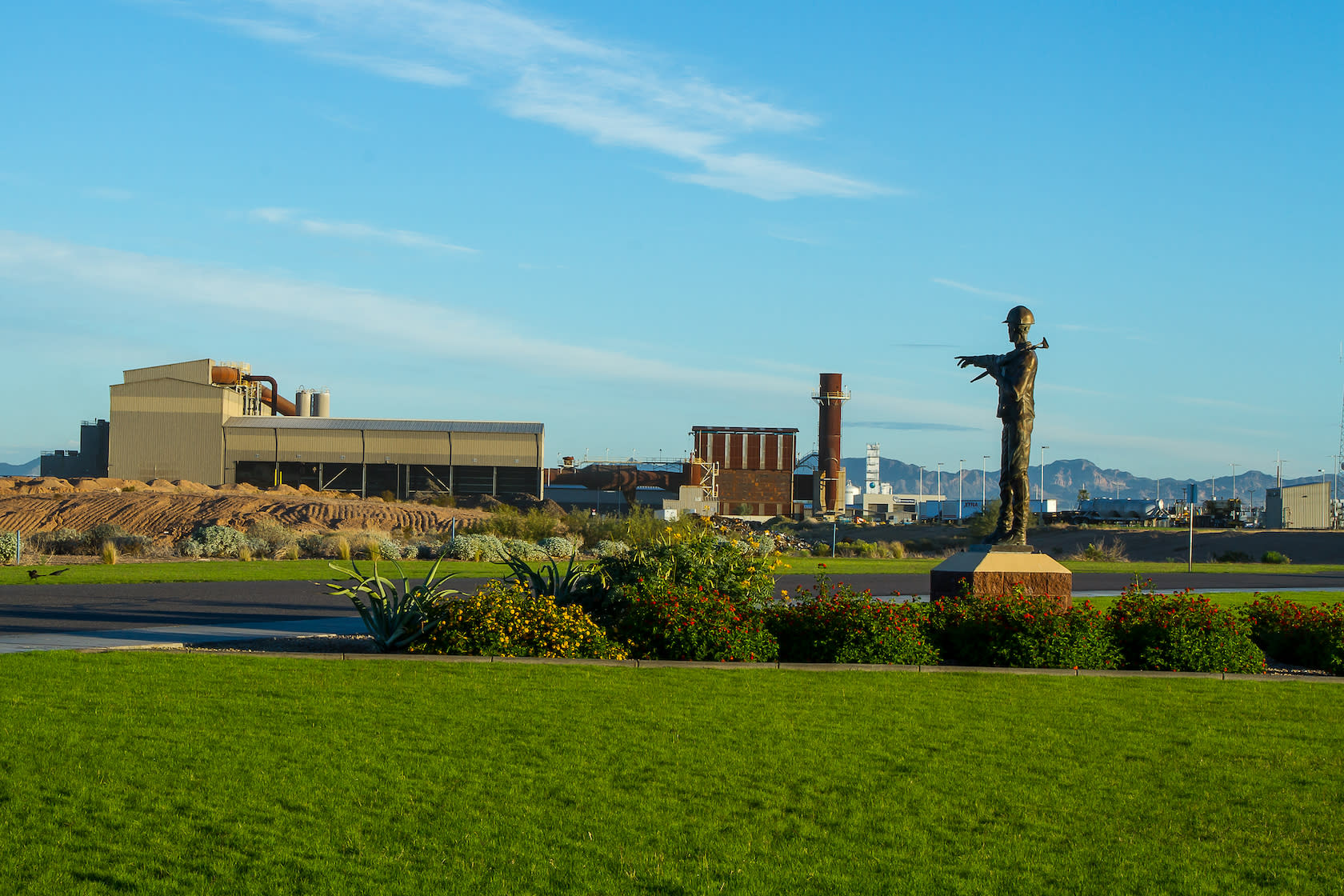 CMC Steel foundry in Mesa, a large beige building with adjacent brick building and stack with a bronze of a steel worker poin