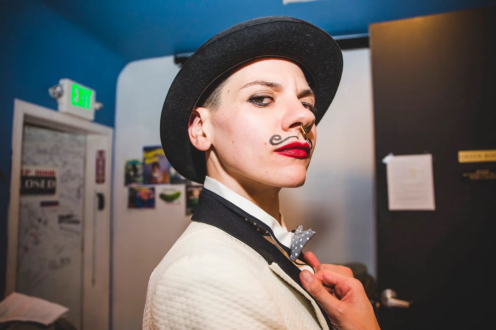 Person with a hat painted on mustache, red lipstick, septum ring, wearing a white suit jacket