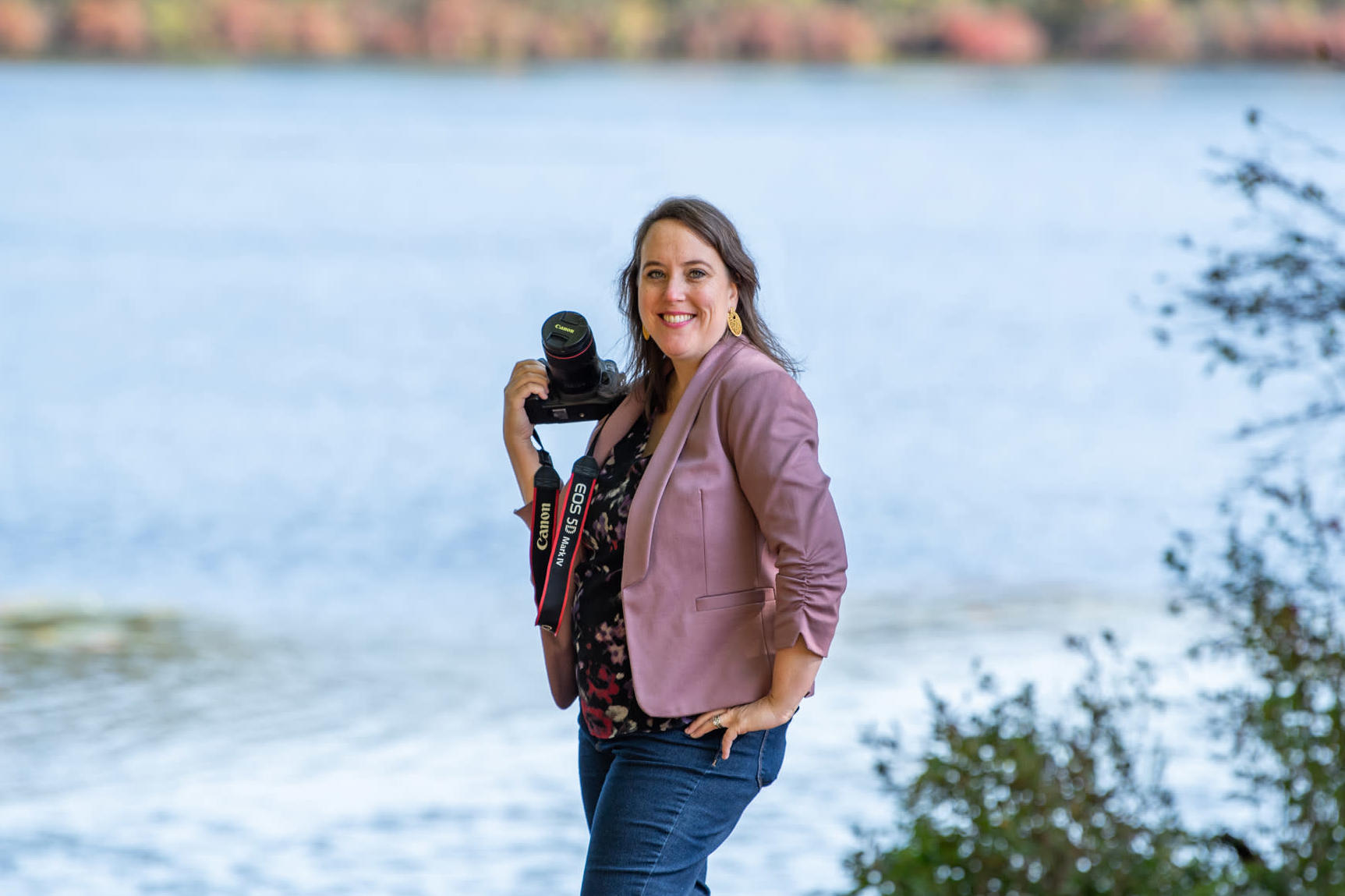 A Caucasian woman holding a Canon DSLR camera by a lake in a purple suit and jeans.