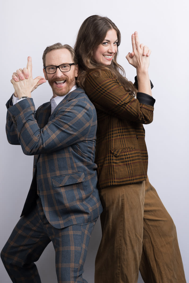 a caucasian man and woman stand back to back, smiling, with finger guns held in front of them.