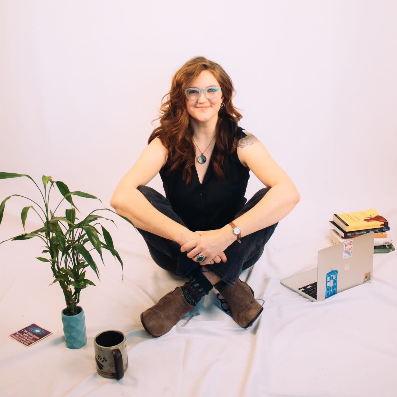Lark smiles- she is sitting on the ground surrounded by a plant, a laptop and design books