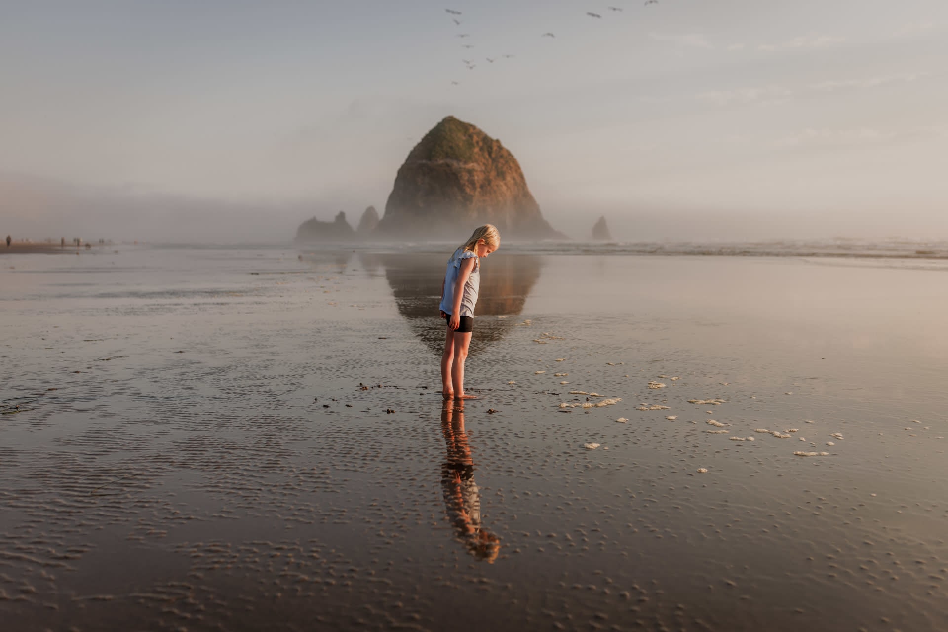 girl standing in front of a rock on the beach and her reflection showing in the water