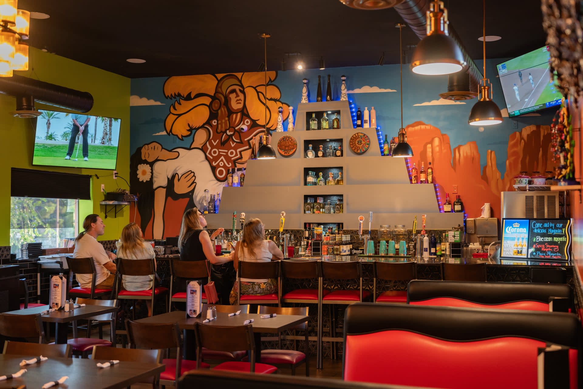 Guests sitting at the bar inside Tortas de Fuego West Sedona with a colorful Aztec-style mural and bottles displayed