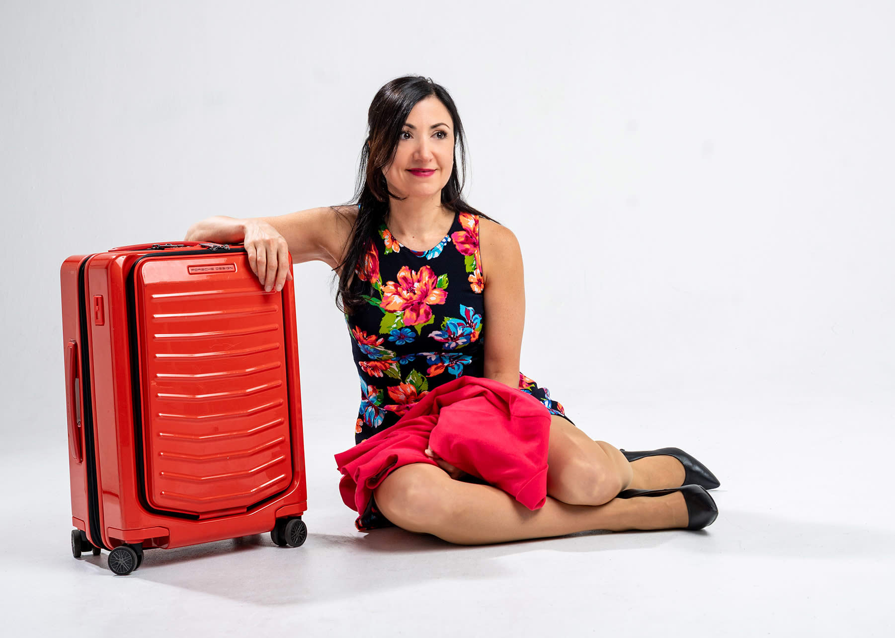 A eoman in a floral dress sits on the ground next to an orange suitcase