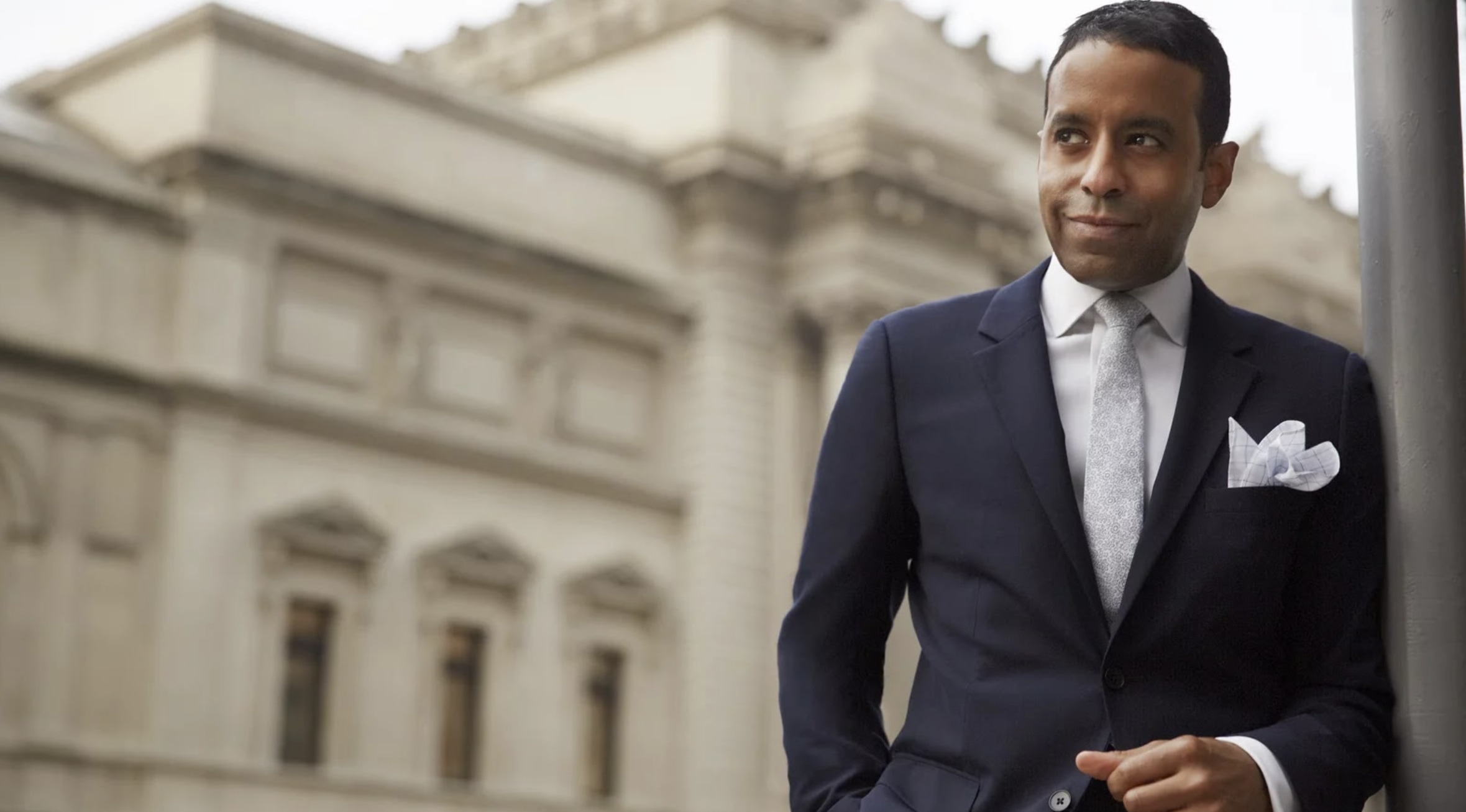 A gentleman in a suit and tie standing on the right corner of the image, smiling as he looks towards the left. A blurry build