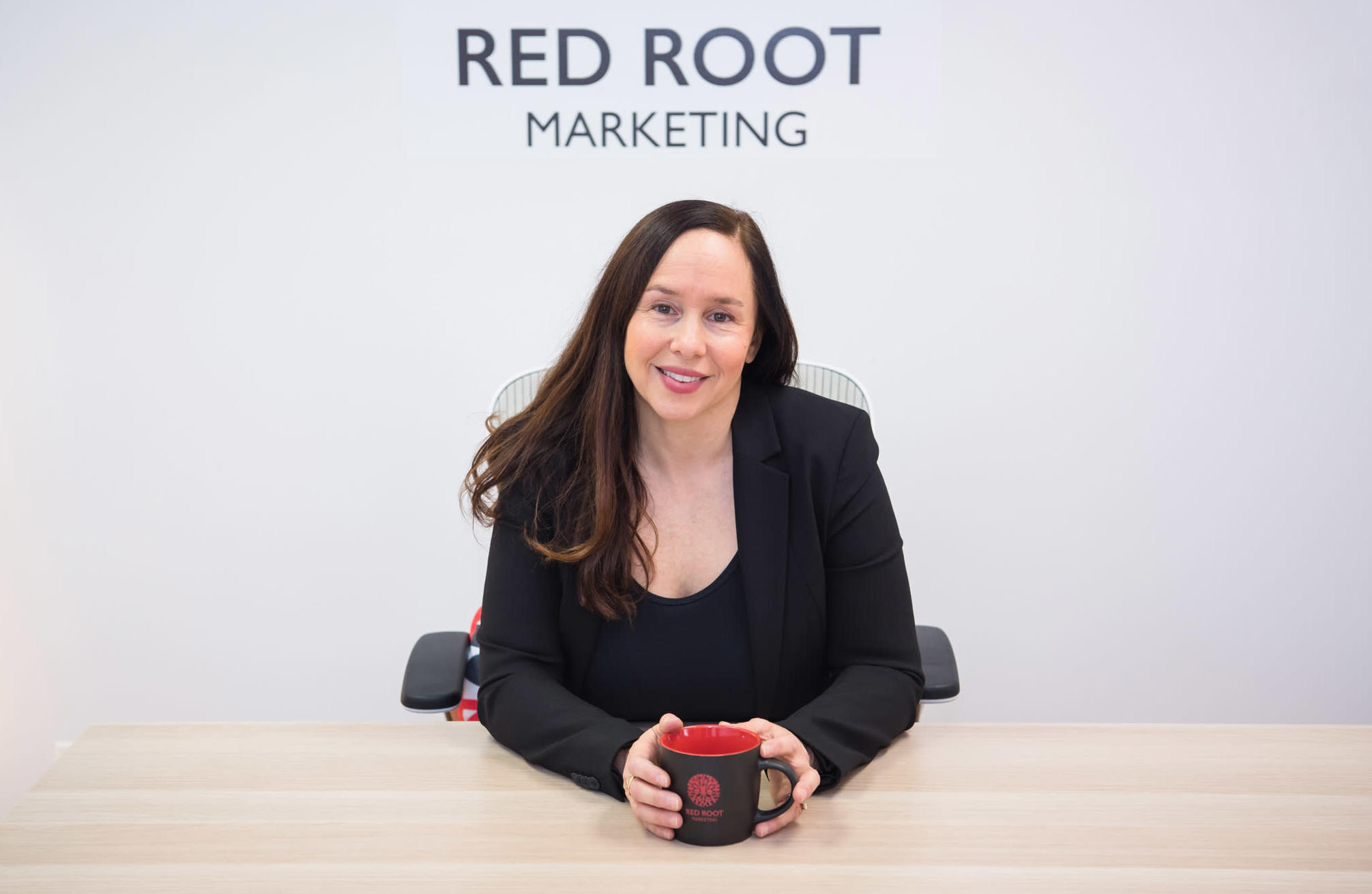 A Caucasian woman sitting down at her office holding a black and red cup in a black suit and shirt.