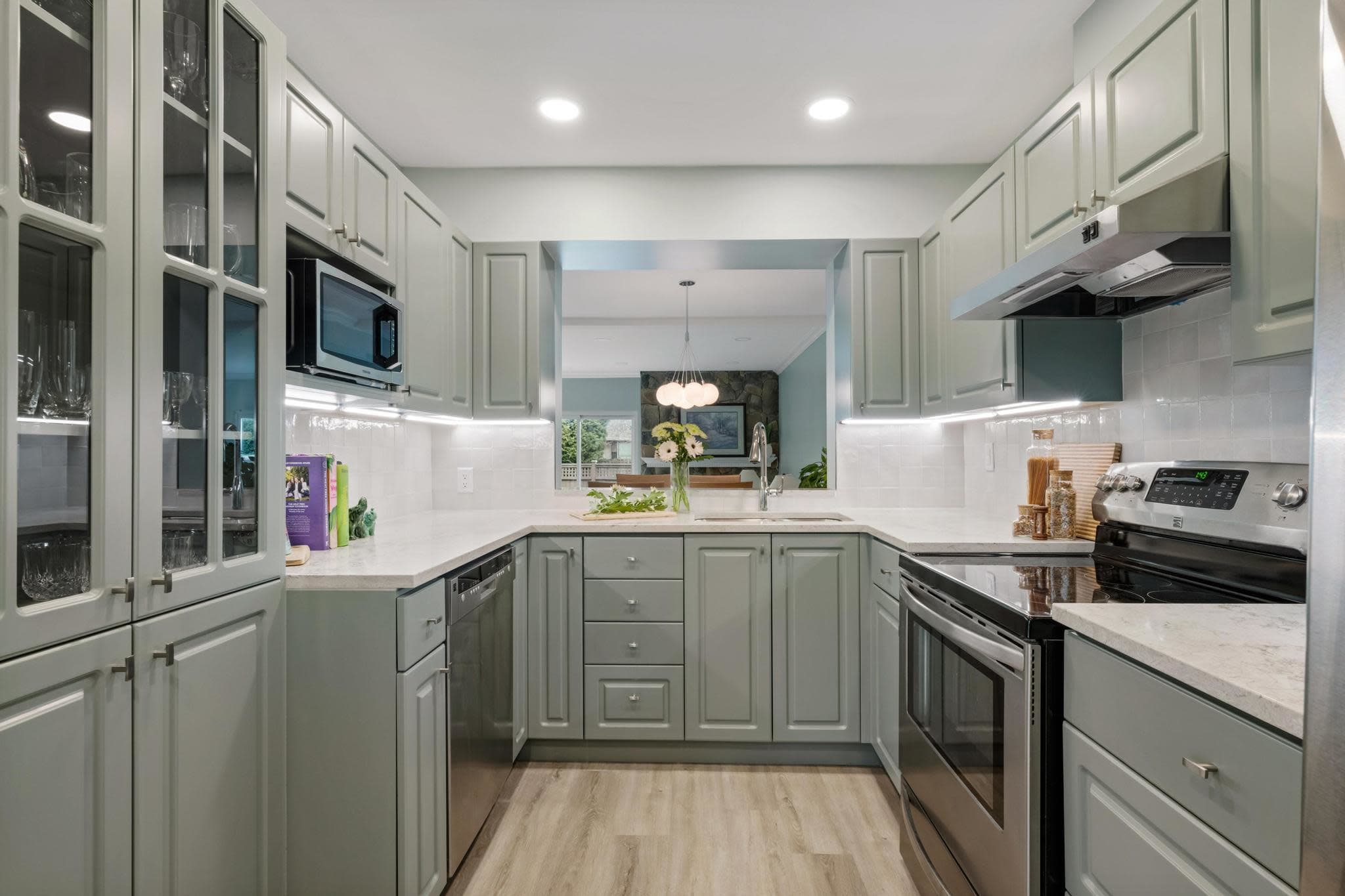 A kitchen. The cabinets are a green-grey, with a white counter and backsplash.