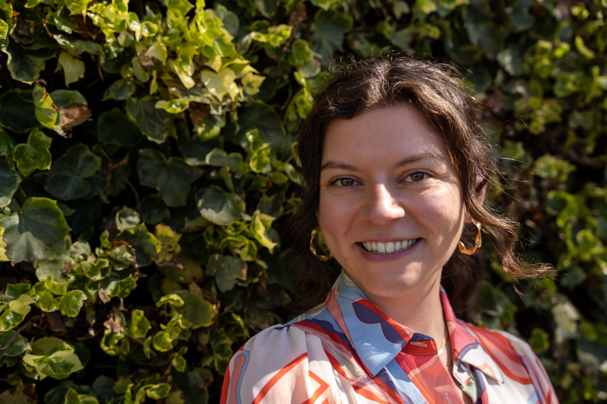 A photo of Melissa Sherry in front of a wall of leaves with a multi-colored dress on