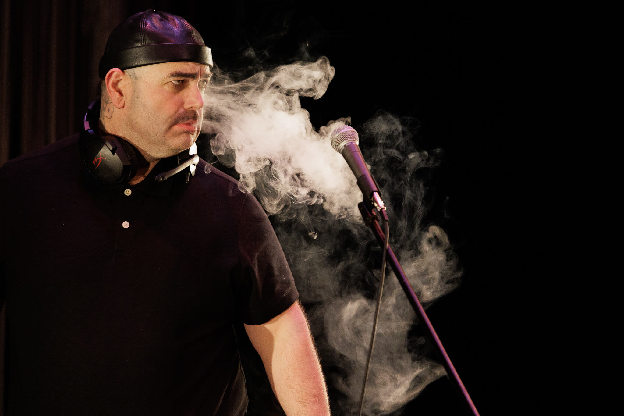 A large person dressed in black has smoke billowing around them, coming out of their face towards a microphone in front of th