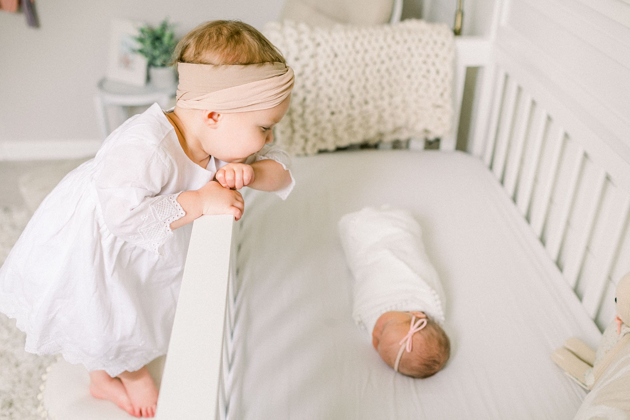 Toddler sister standing on stool leaning over crib to look at her swaddled baby sister asleep.