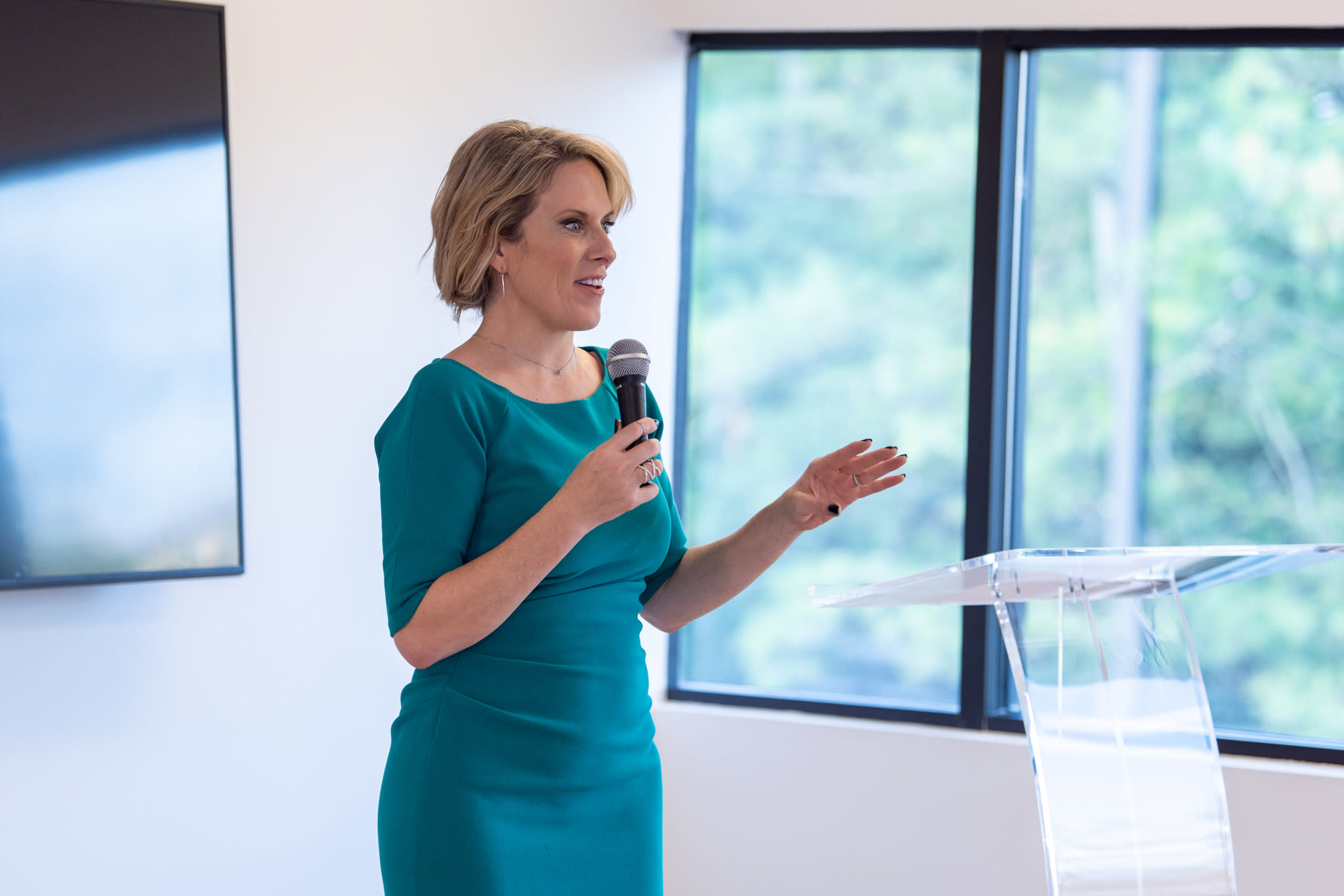 A Caucasian woman in a bluish green dress speaking at a podium holding a mic.
