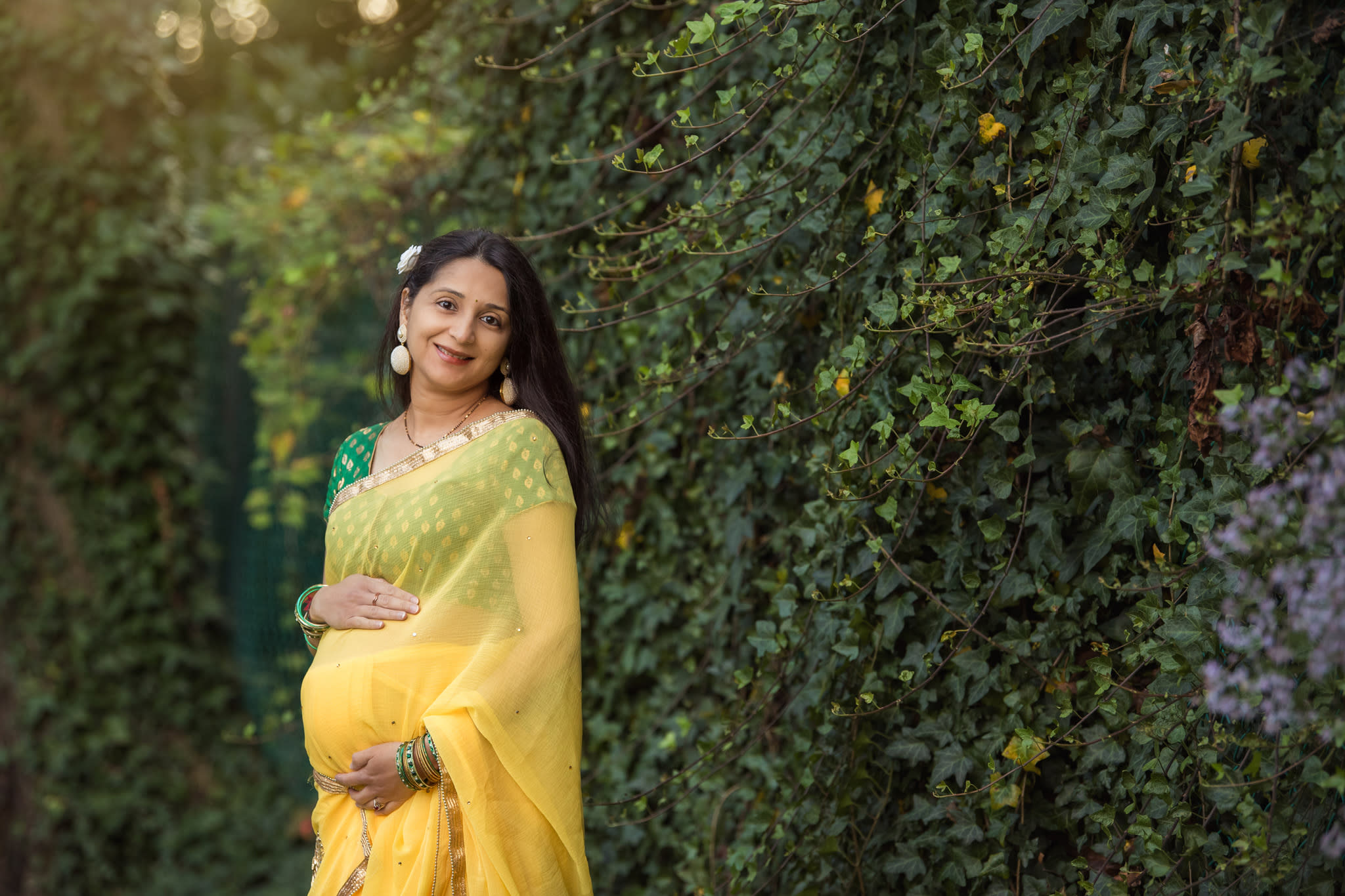 An Indian pregnant woman wearing a yellow and green sari smiling with green vines in the background.