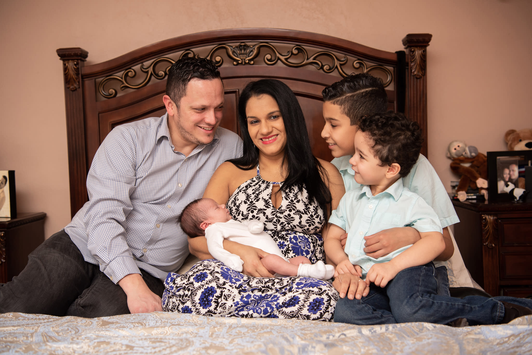 A Latina family of 5 sitting on the bed with their newborn son at their newborn photography session.