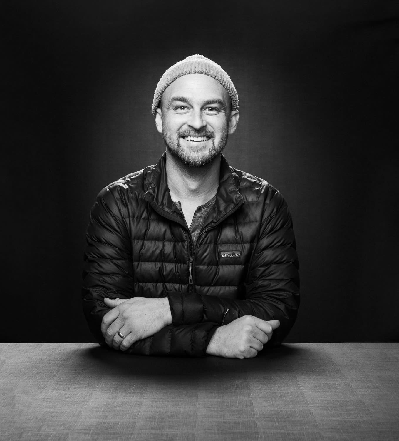 Studio Owner shown from the table up in black and white. He has a knit cap on his head and a puffy jacket.