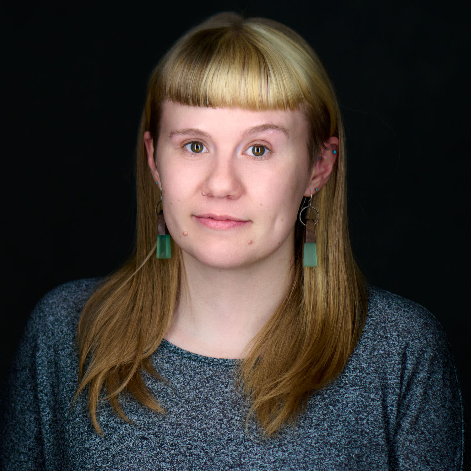 A person with straight, light brown hair and blunt bangs, wearing green rectangular earrings and a gray top, poses against a