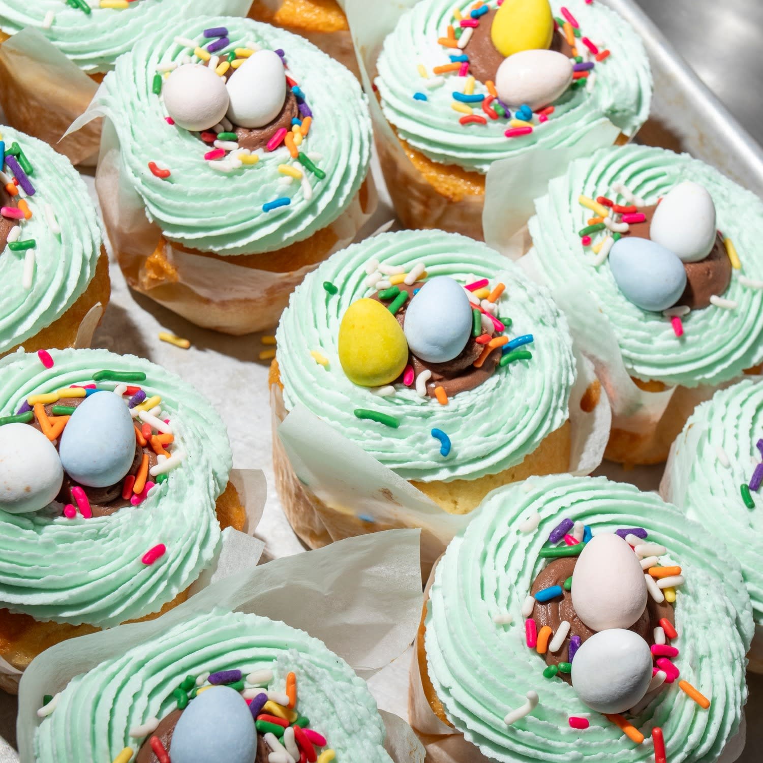 Cupcakes topped with light green frosting, colorful sprinkles, and candy eggs, resembling small nests, arranged on a tray.