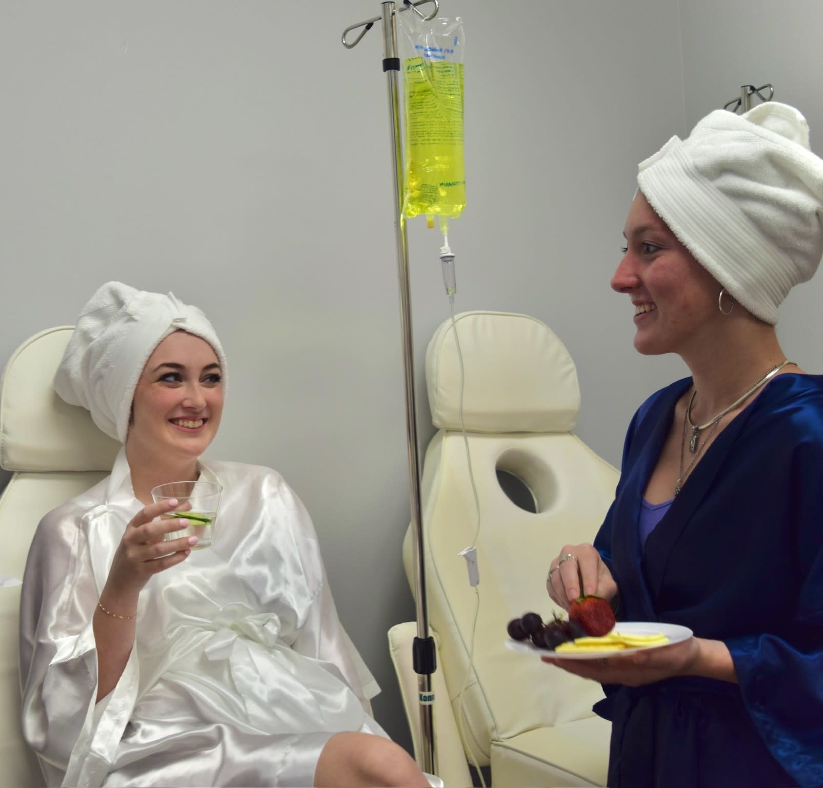Two women in spa robes at a medical spa—one receiving an IV treatment while the other enjoys appetizers.