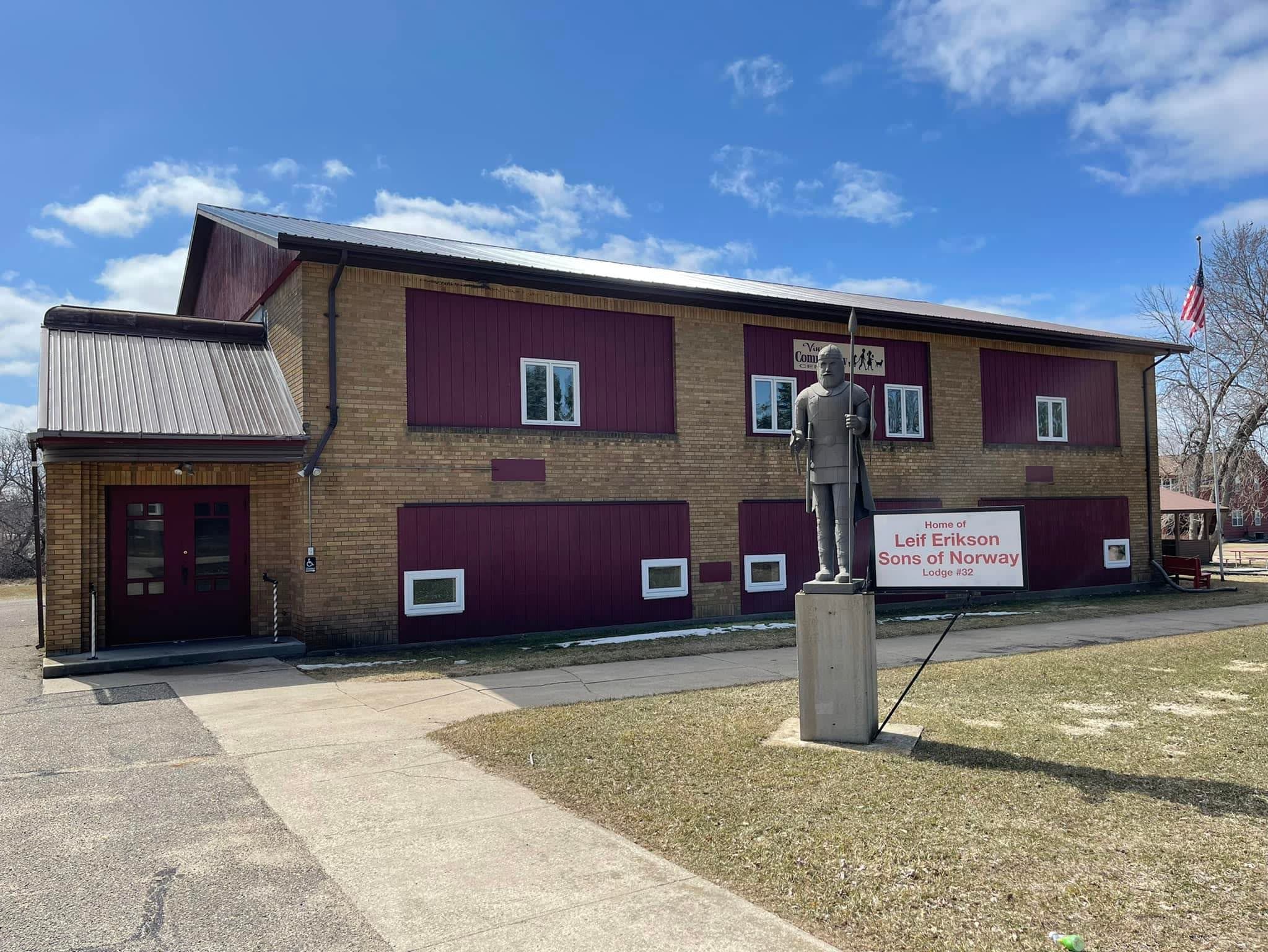 Brick building with metal roof, burgundy wood accents and white windows. Blue sky, white wispy clouds, American flag on pole