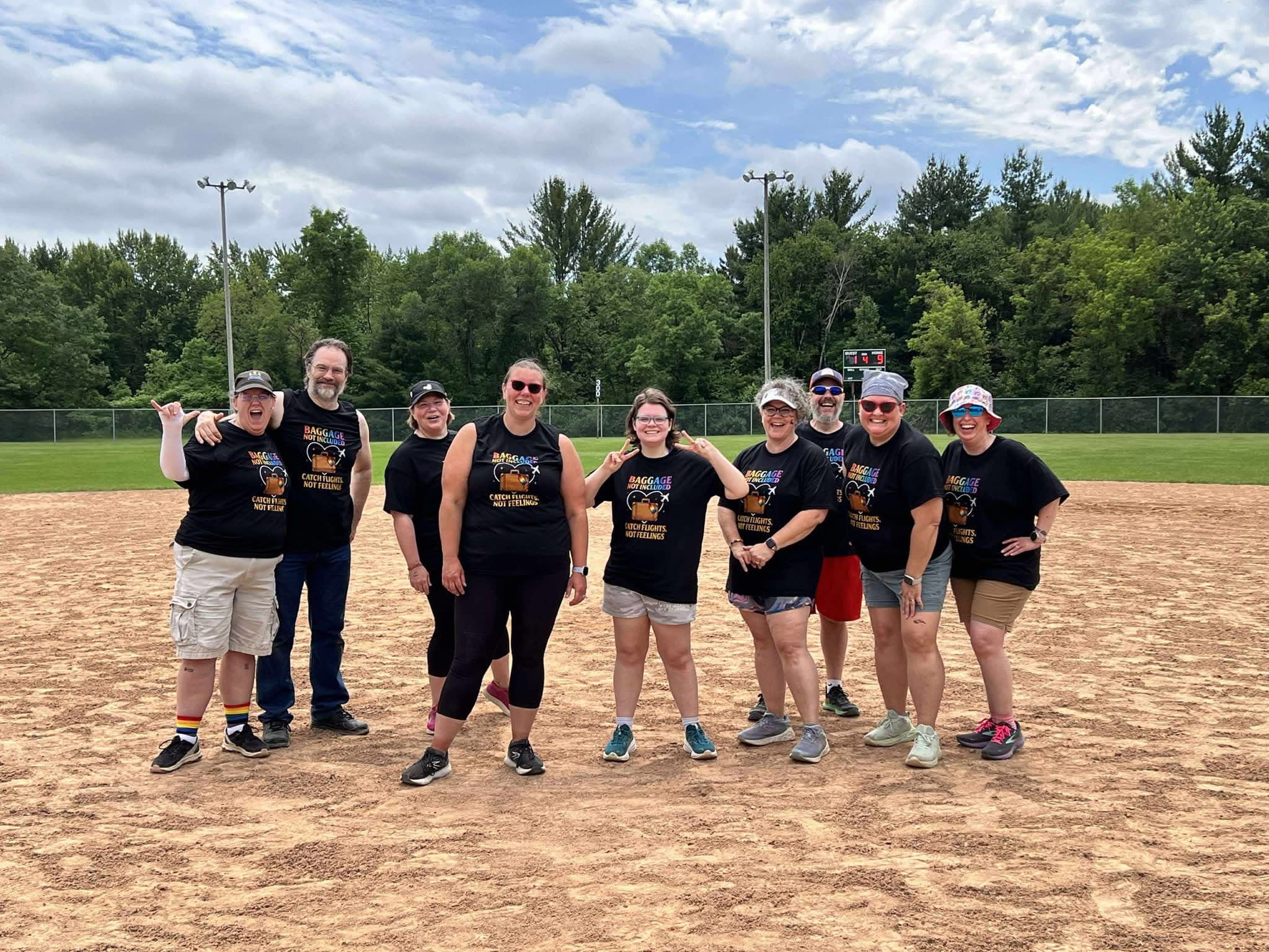 A group of adults wearing matching “Baggage Not Included” shirts stands smiling on a kickball field during a June 2025 fundra