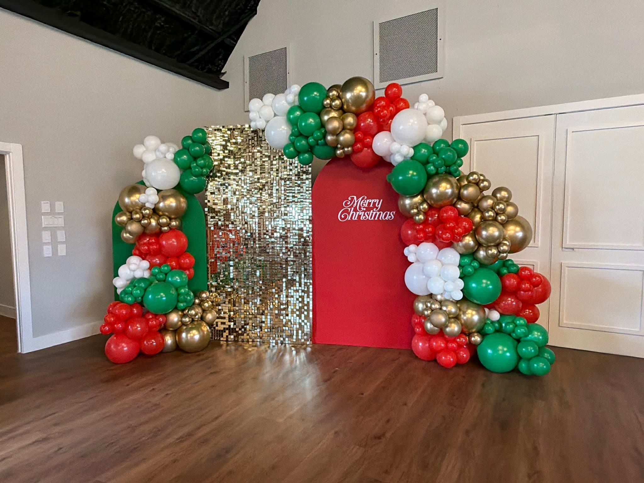 Christmas balloon arch installation with red, green, white, and gold balloons framing three backdrop panels, including a gold
