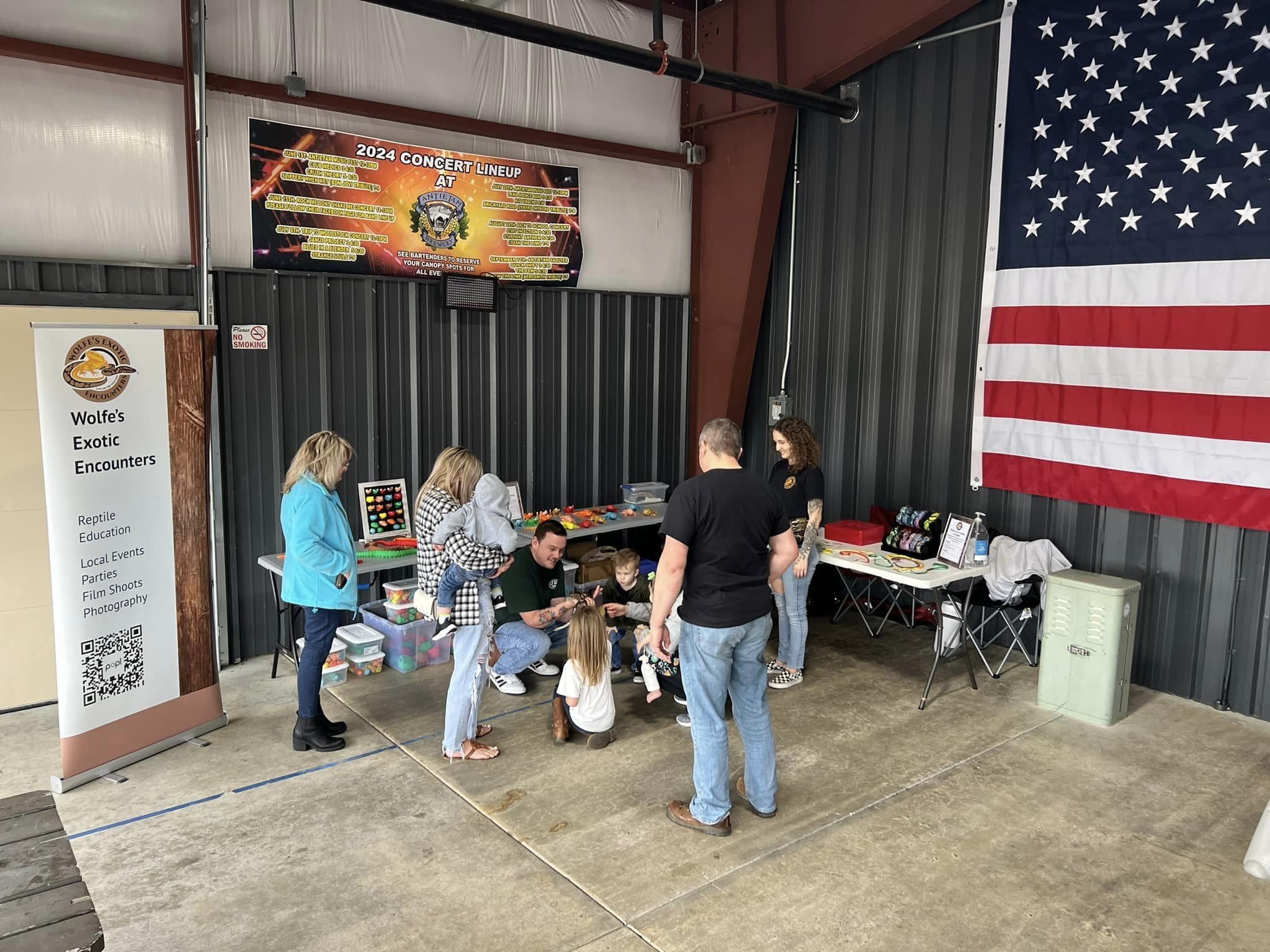 Indoor booth with 'Wolfe's Exotic Encounters' banner, people interacting with reptiles, tables with displays.