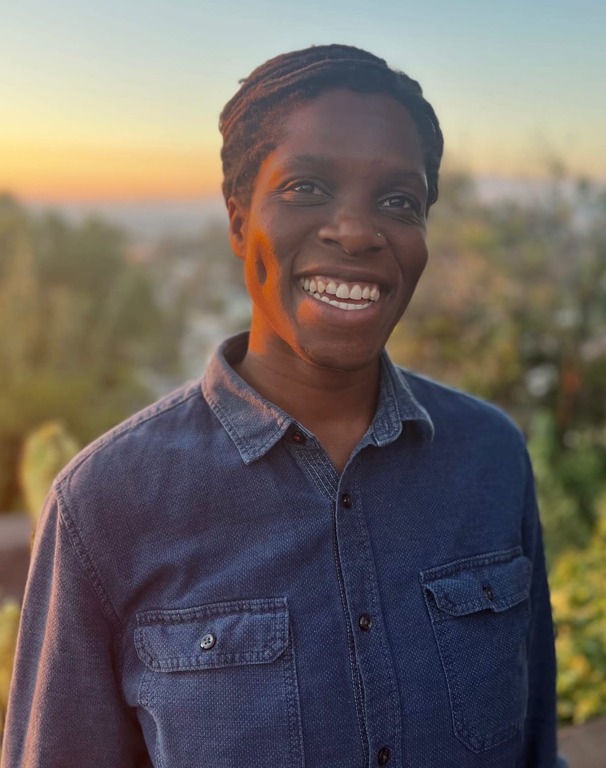 Chika, smiling, in a blue button-down shirt, with green plants and the blues and pinks of the Los Angeles sunset as backdrop