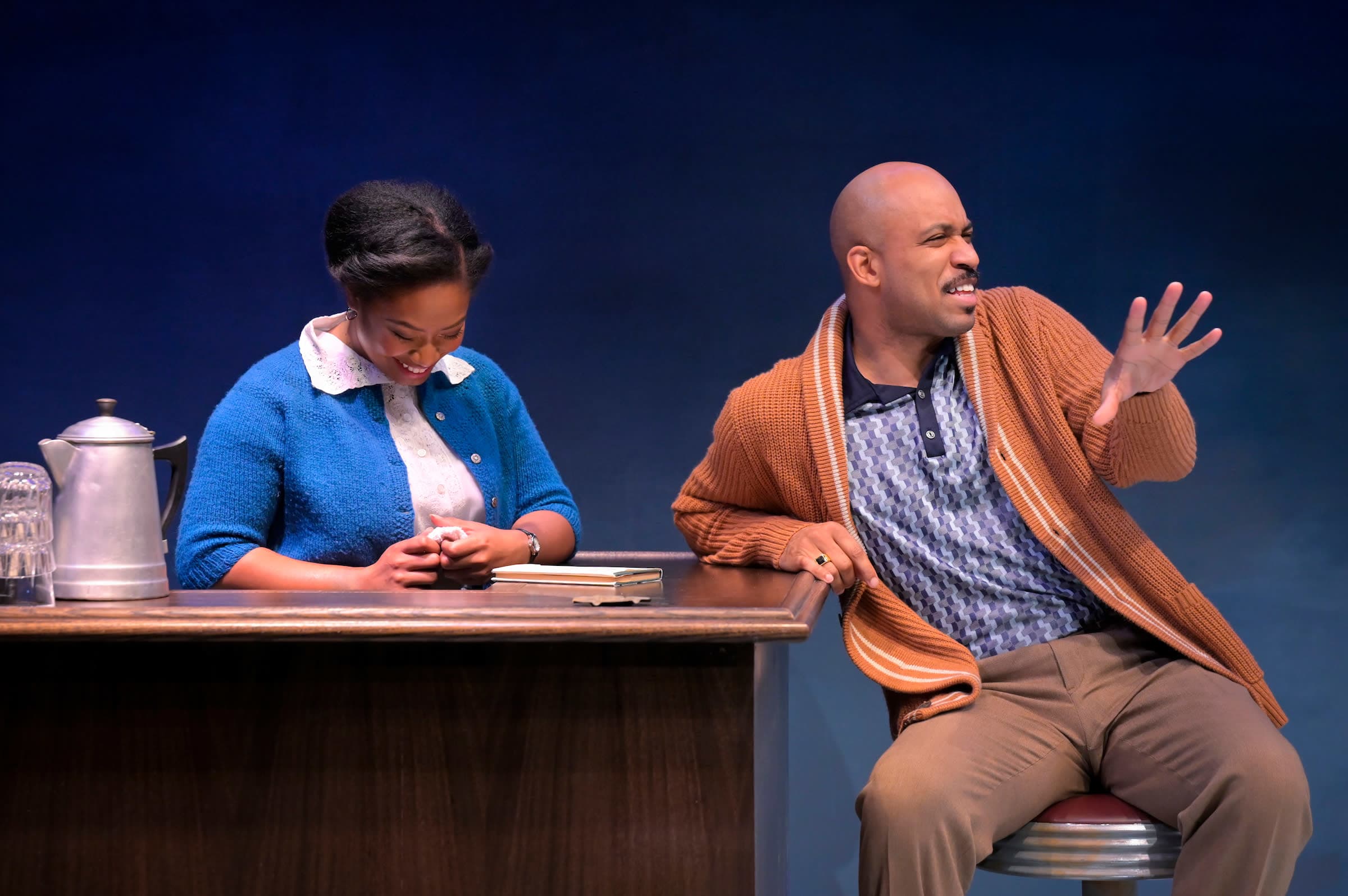 Black woman in a blue sweater standing behind a bar laughing, light skinned black man in a peach cardigan sits on a bar stool