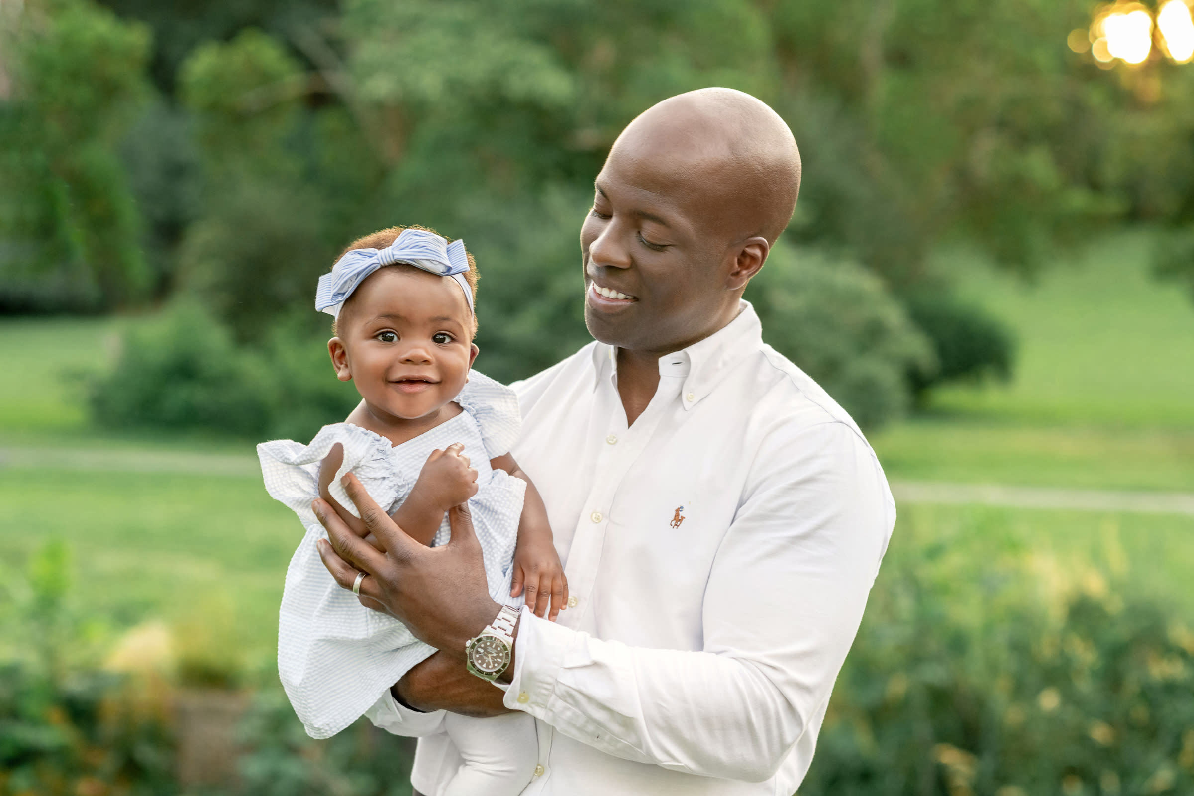 African American dad in white shirt holding and lovingly looking at 6 month old daughter outside in park at sunset