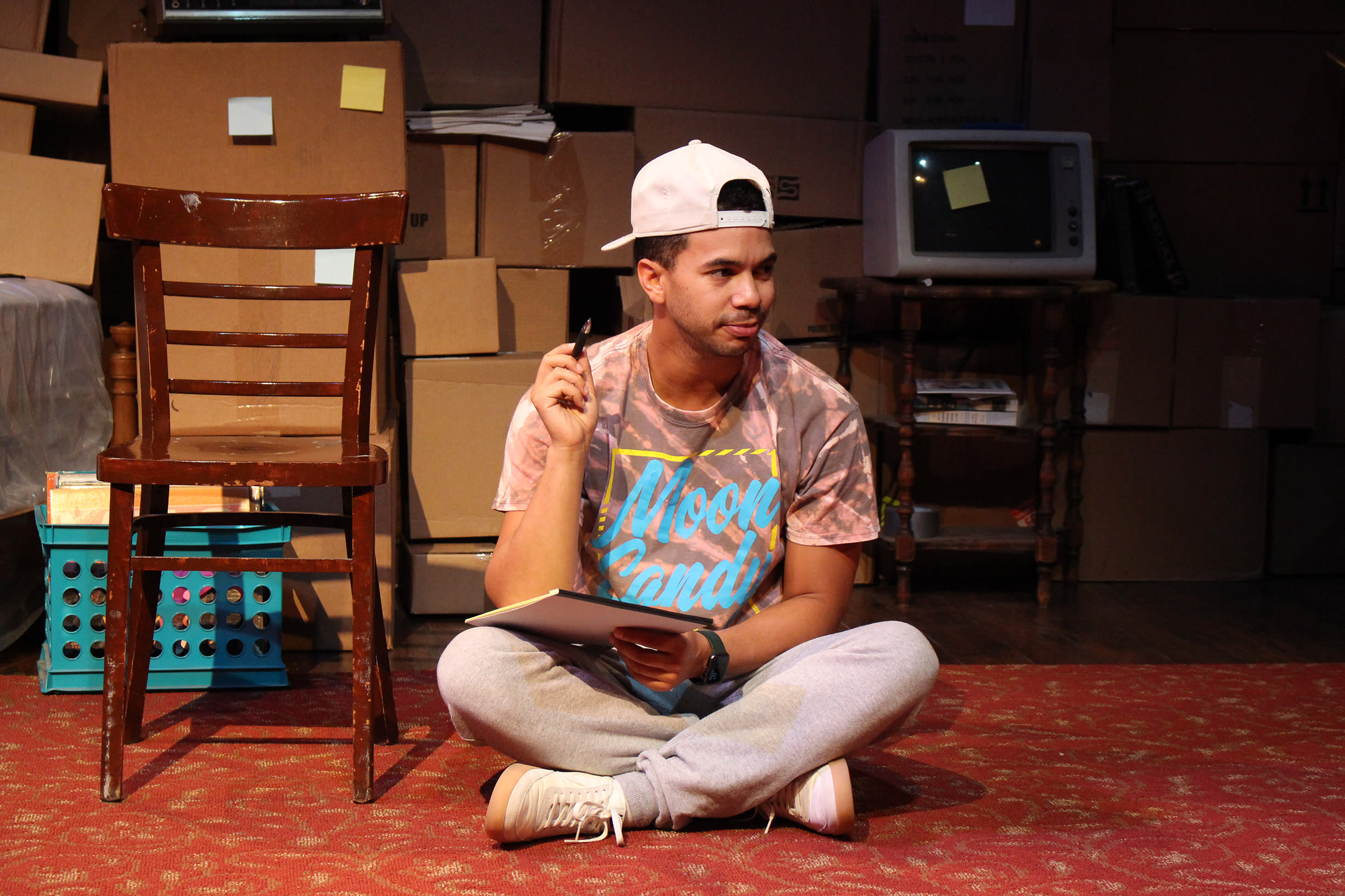 An actor sits crossed legged on the ground holding a pad of paper and pen.