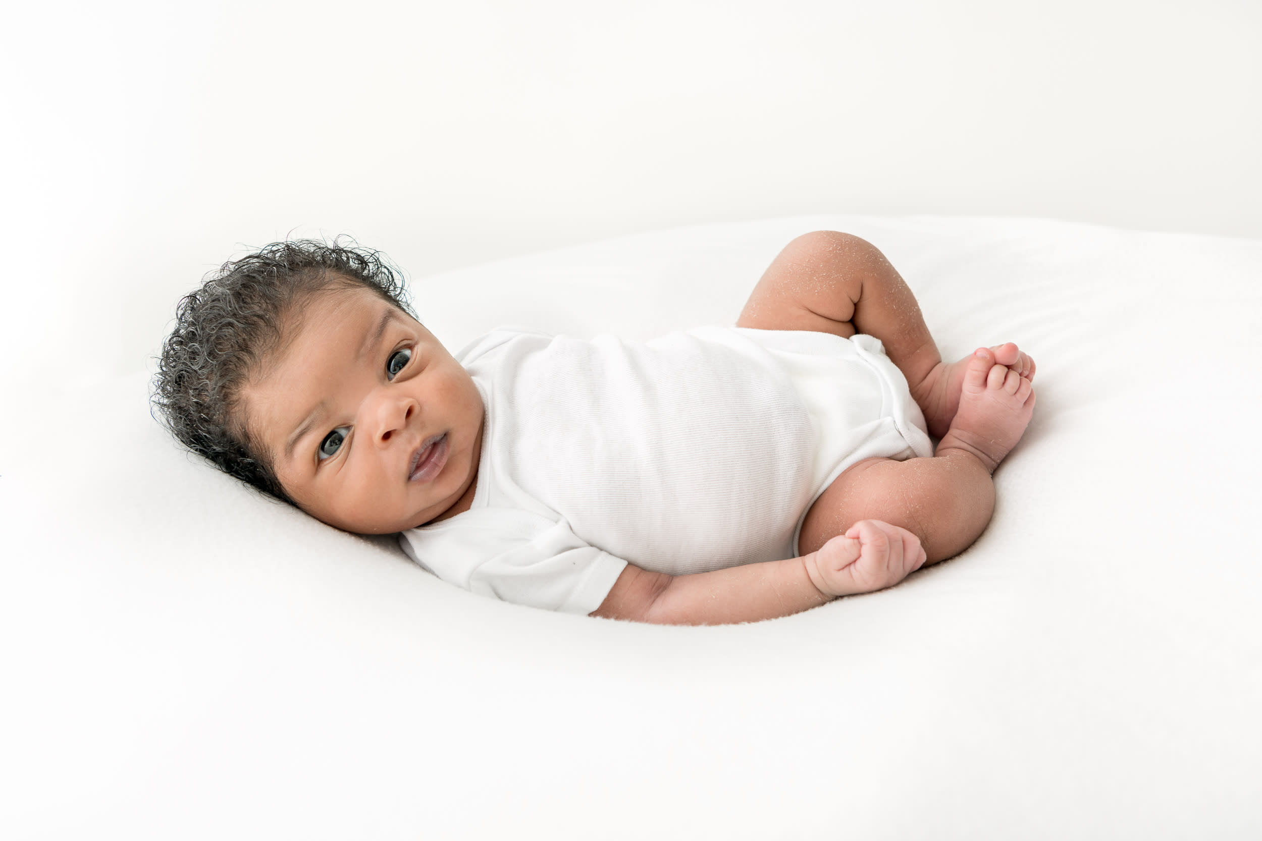 Newborn baby boy with curly hair and medium skin in white onesie curled up on white beanbag staring into the camera