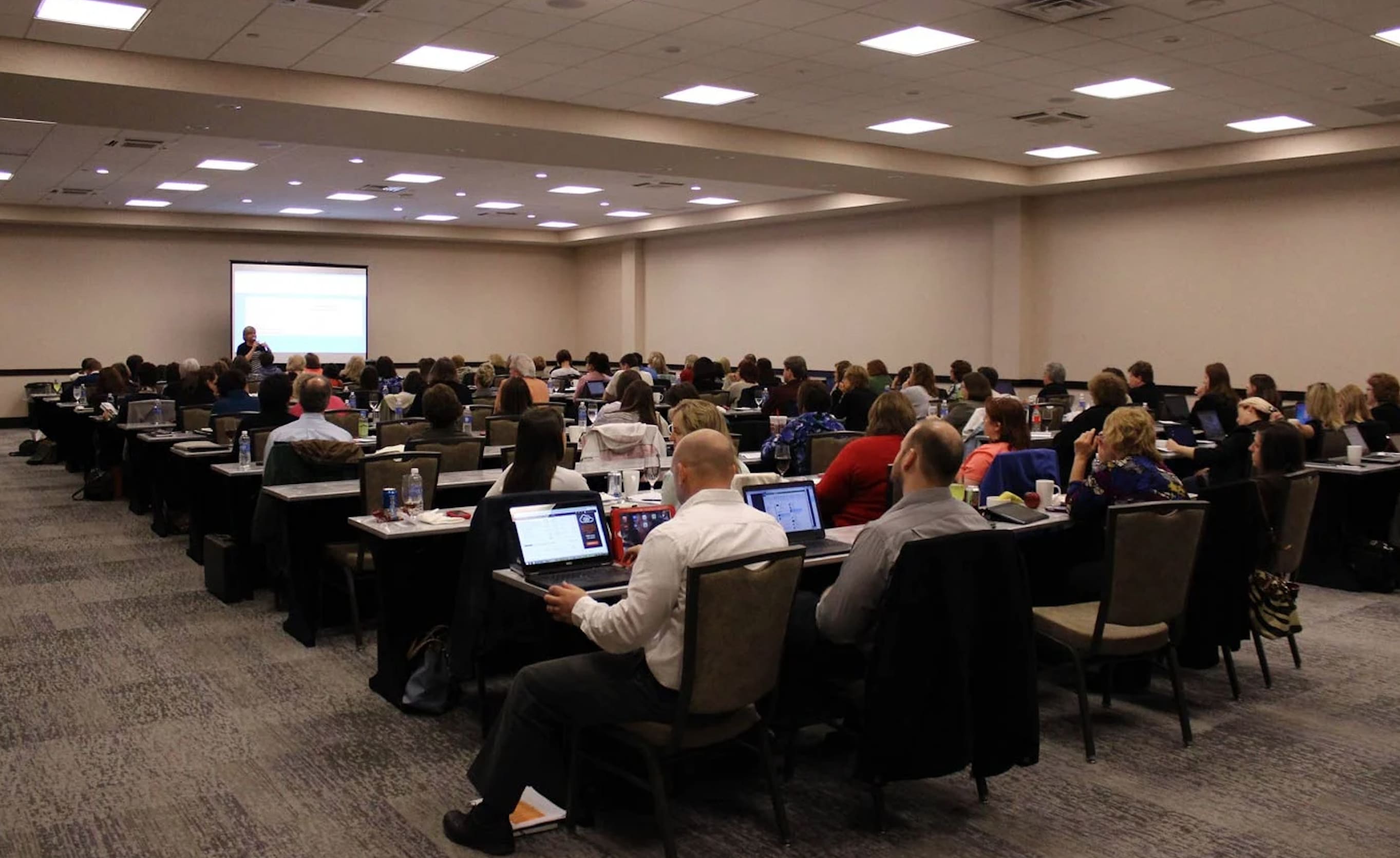 Conference Attendees in large conference room, seated in rows with tables, laptops, and notebooks in front of them.